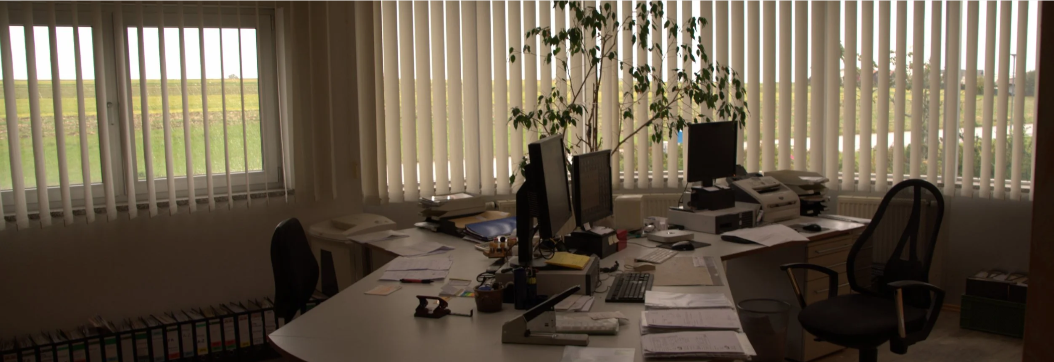 Ein chaotischer Büroarbeitsplatz mit mehreren Computerbildschirmen, Papieren, Aktenordnern und Büromaterialien auf einem großen Schreibtisch vor großen Fenstern mit vertikalen Jalousien, durch die Tageslicht scheint, und einem Pflanzen im Hintergrund.