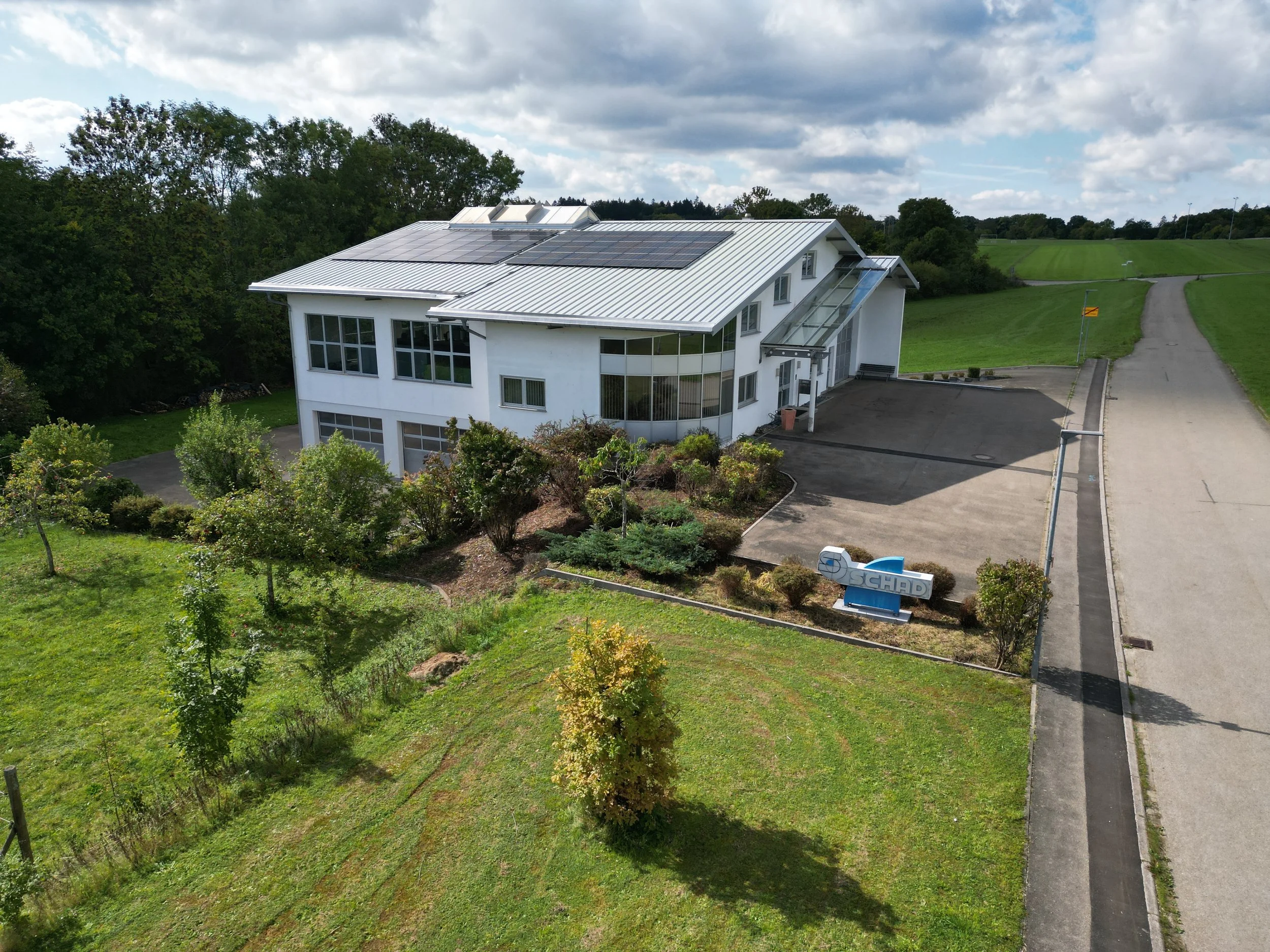 Ein modernes Bürogebäude mit Solarzellen auf dem Dach, umgeben von Grünflächen und Bäumen, mit einem Parkplatz im Vordergrund.