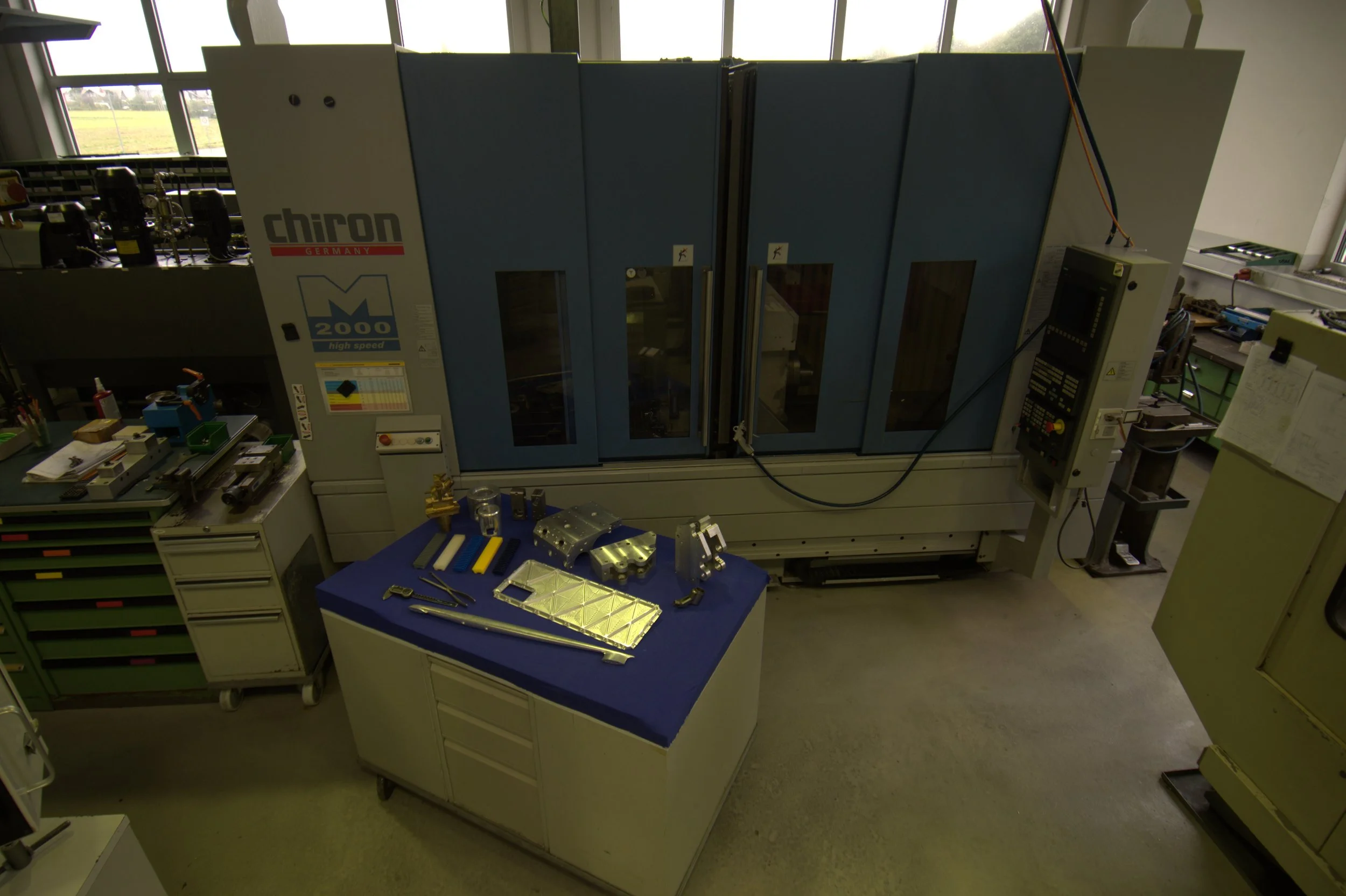 Industrieller Maschinenraum mit verschiedenen Werkzeugen auf einem Tisch und einer großen CNC-Maschine (Chiron 2000 high speed) im Hintergrund.
