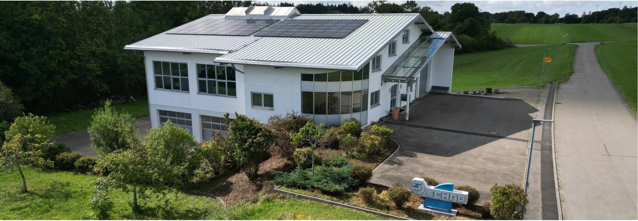 Modern, weißes Gebäude mit großen Fenstern und Solarpanelen auf dem Dach, umgeben von grüner Landschaft und Bäumen.