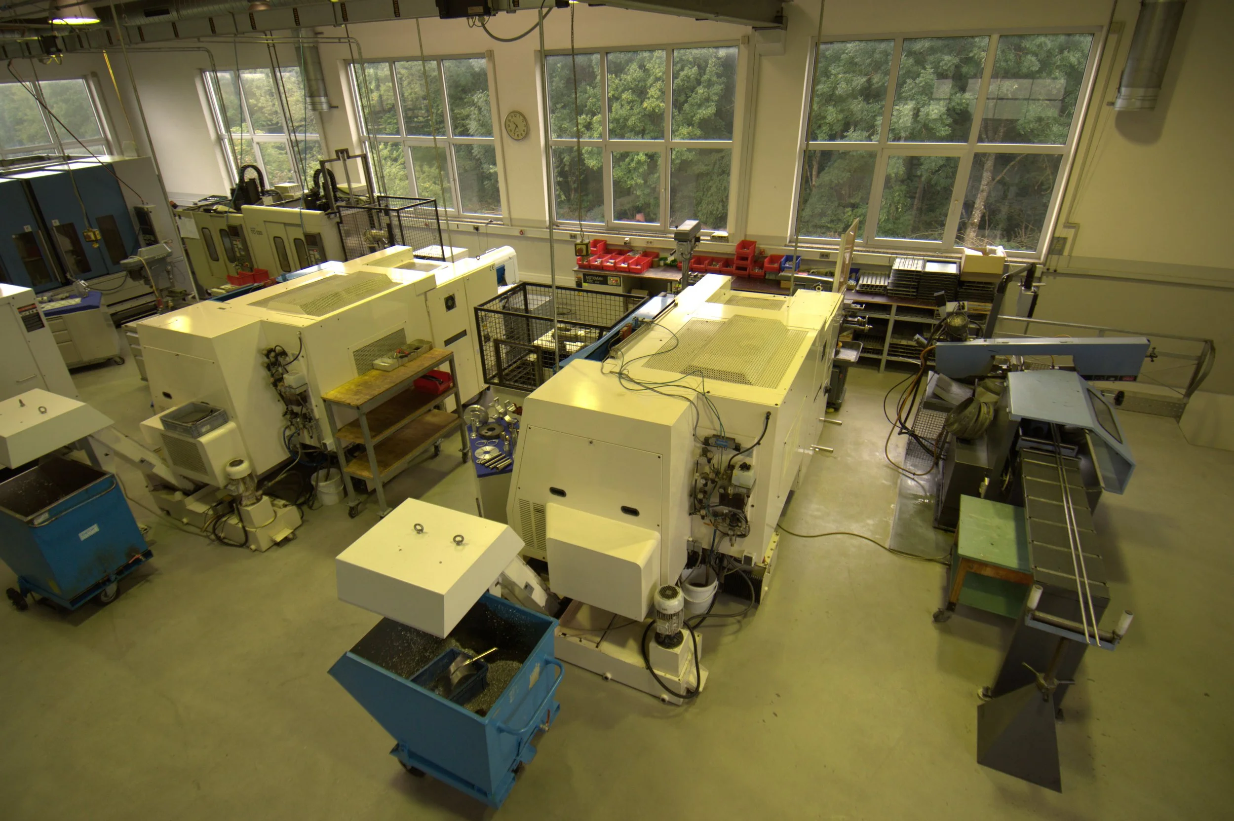 Laborraum mit großen, weißen Maschinen und Werkzeugen, gelbe und blaue Behälter, große Fenster mit Blick auf Bäume im Hintergrund.