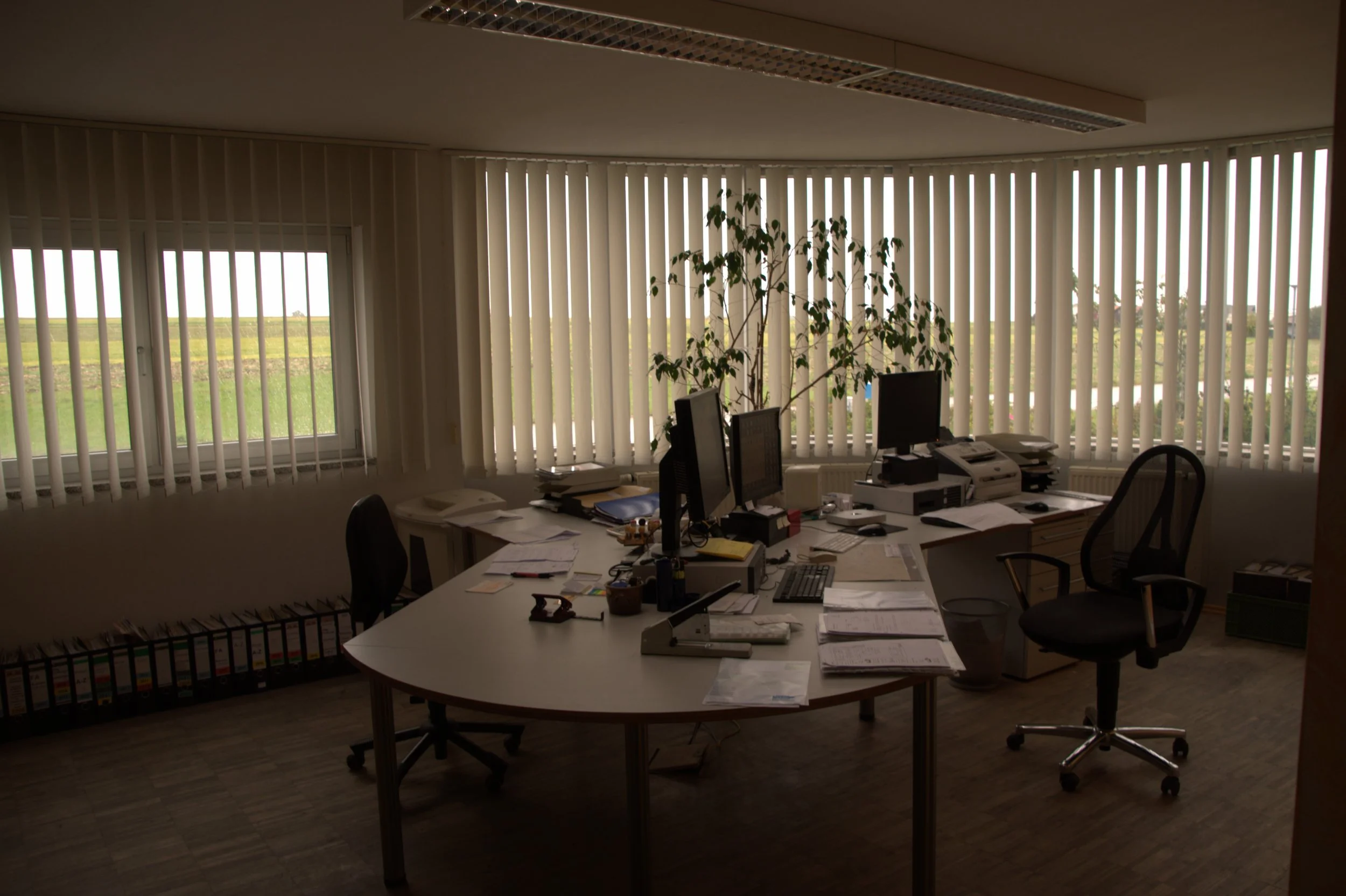 Ein unordentlicher Büroarbeitsplatz mit mehreren Computerbildschirmen, Papieren, Büromaterialien und zwei Bürostühlen vor großen Fenstern mit vertikalen Jalousien, Blick auf eine grüne Landschaft im Hintergrund.