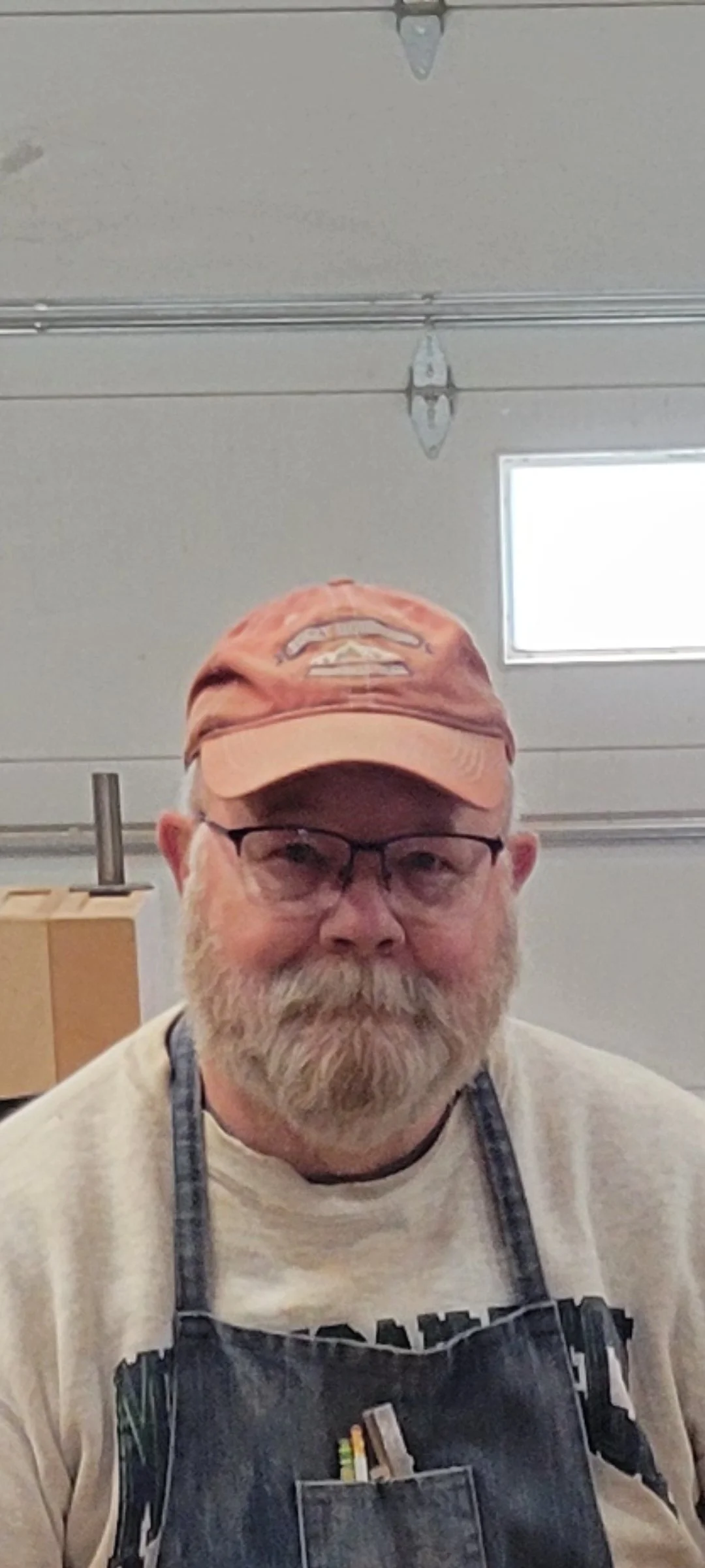 An older man with a beard and glasses wearing a baseball cap and apron, standing indoors near a garage door.