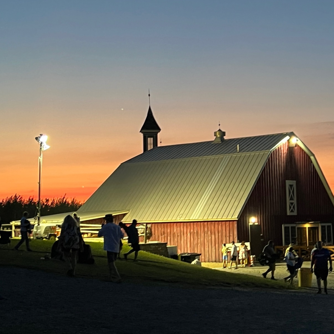 People walking and gathering outside a large red barn with a metal roof at sunset, with a small steeple on the barn and tall lights illuminating the area.