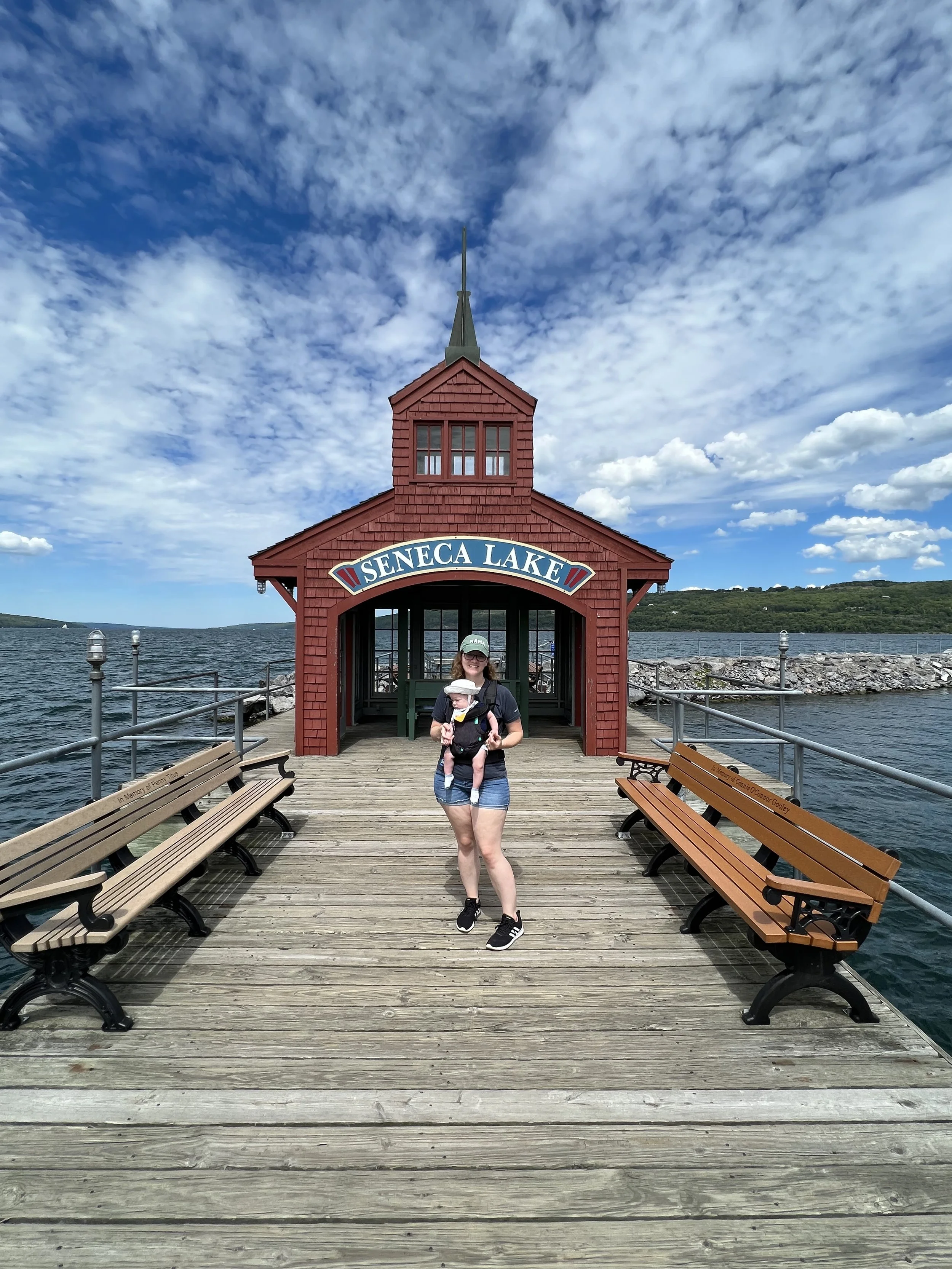 Woman and child on a wooden dock in front of a red boathouse with a sign that reads 'Seneca Lake', overlooking a lake under a partly cloudy sky.