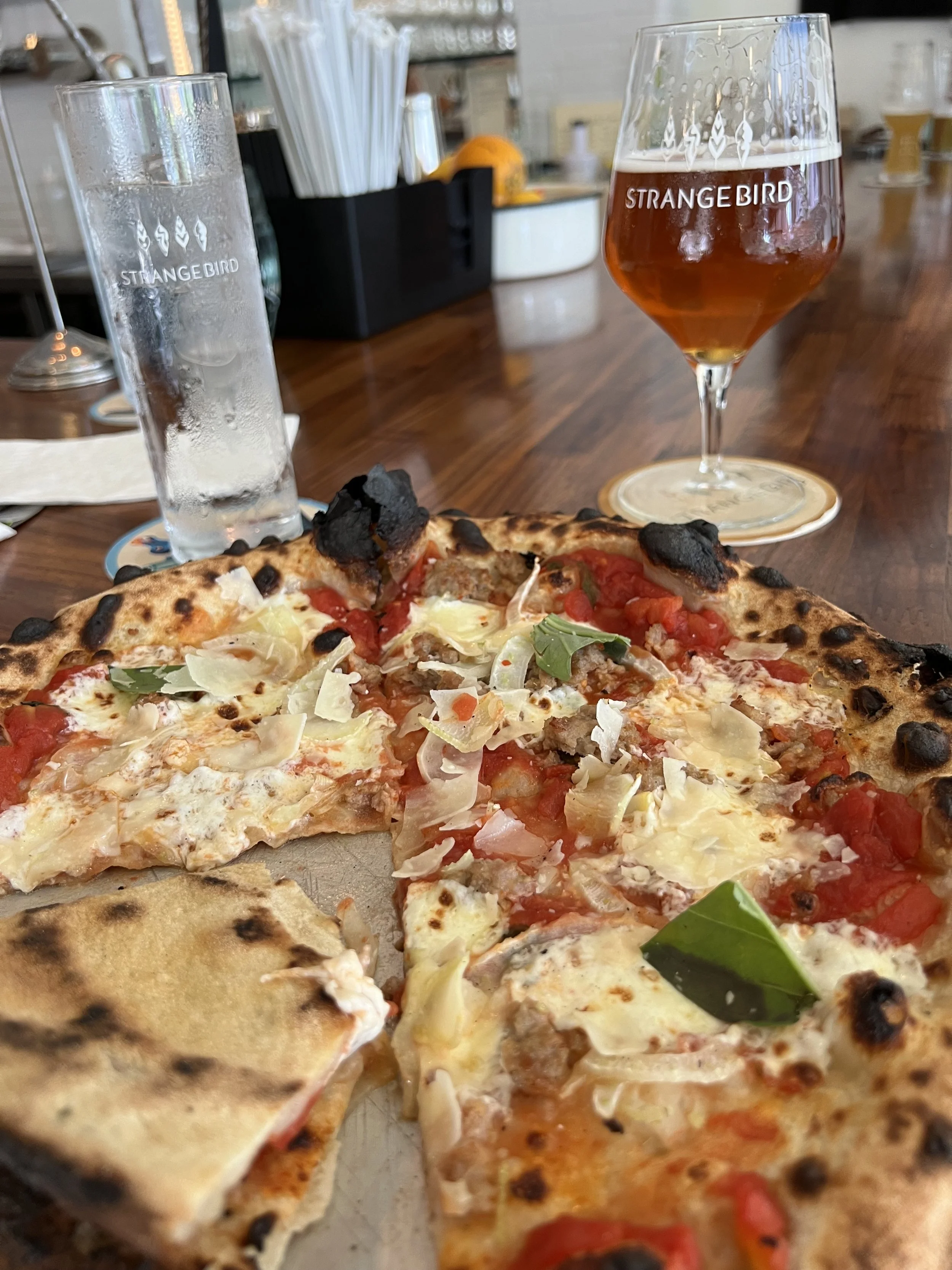 A pizza with cheese, tomatoes, and basil on a wooden table. Two drinks, one clear and one amber-colored, are in the background at a restaurant or bar.