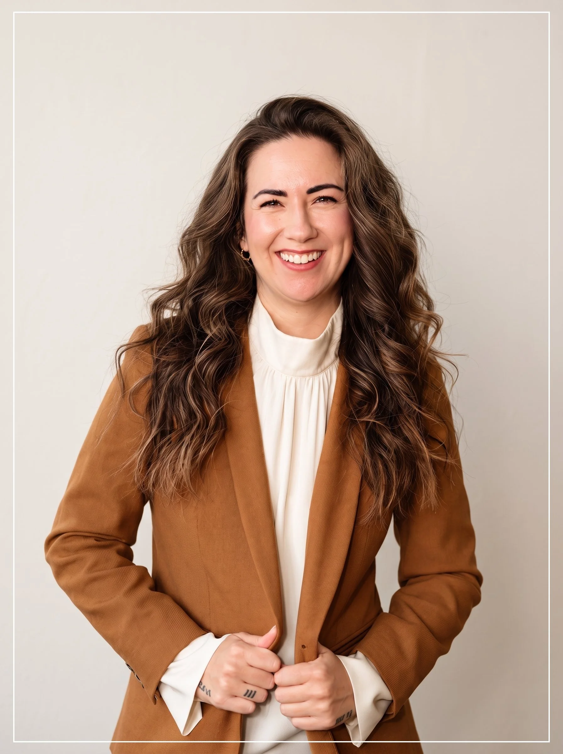 A woman with long, wavy brown hair wearing a brown blazer over a cream blouse, smiling with her eyes slightly squinted, standing against a plain light gray background.