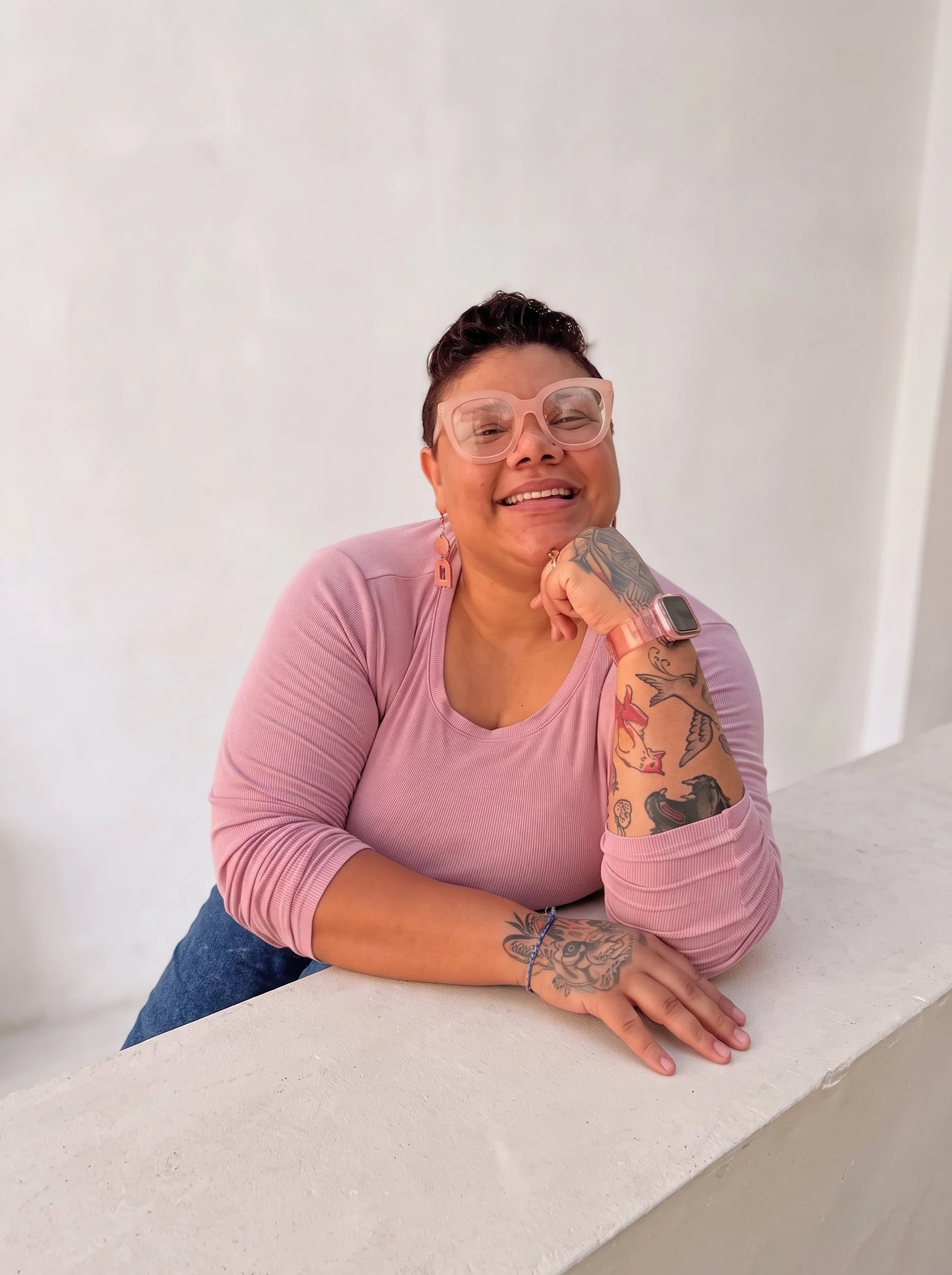 Smiling woman with glasses and tattoos sitting at a white table against a plain white wall