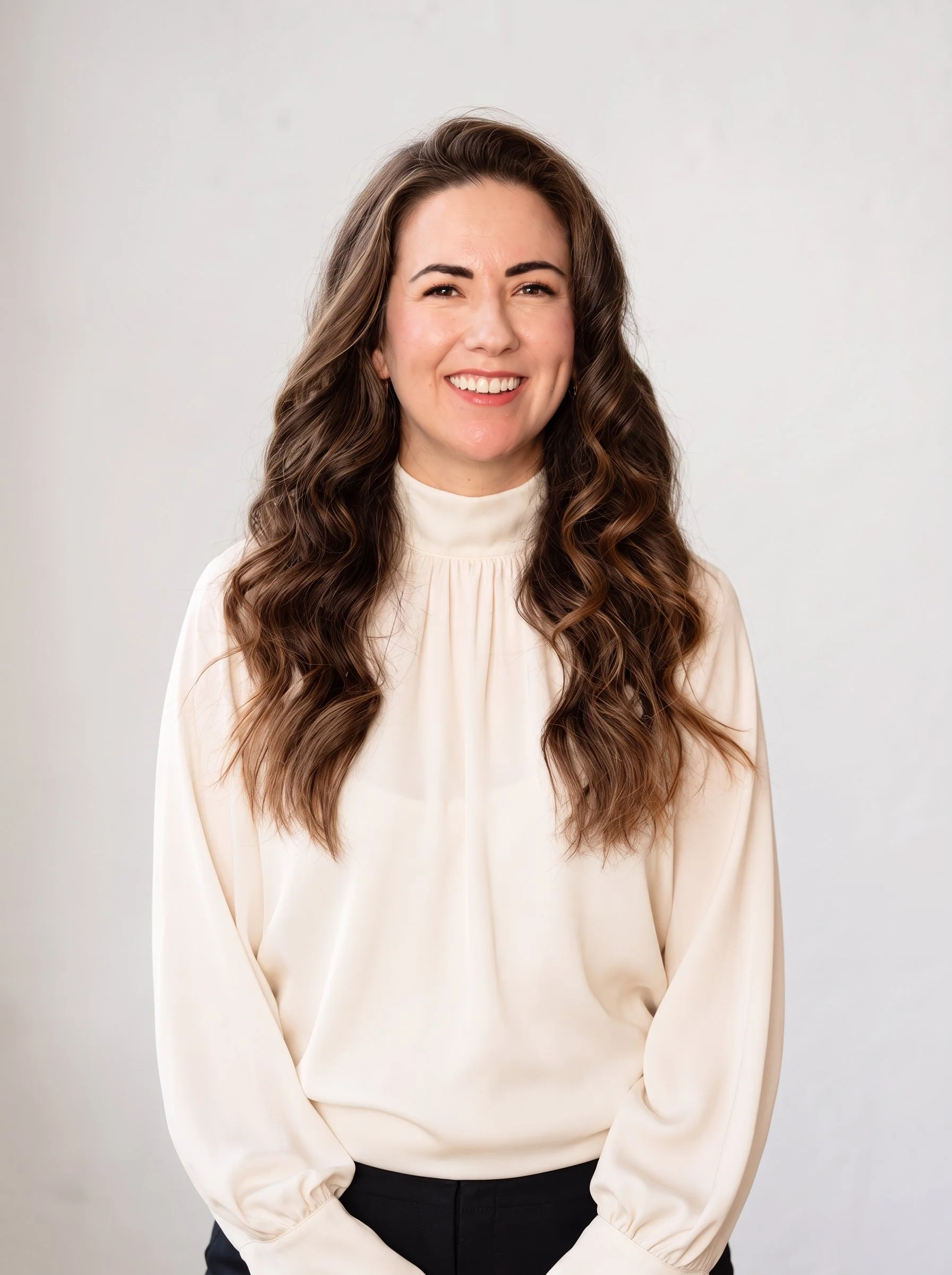A woman with long, wavy brown hair wearing a cream-colored blouse with a high collar, standing against a plain light background, smiling at the camera.
