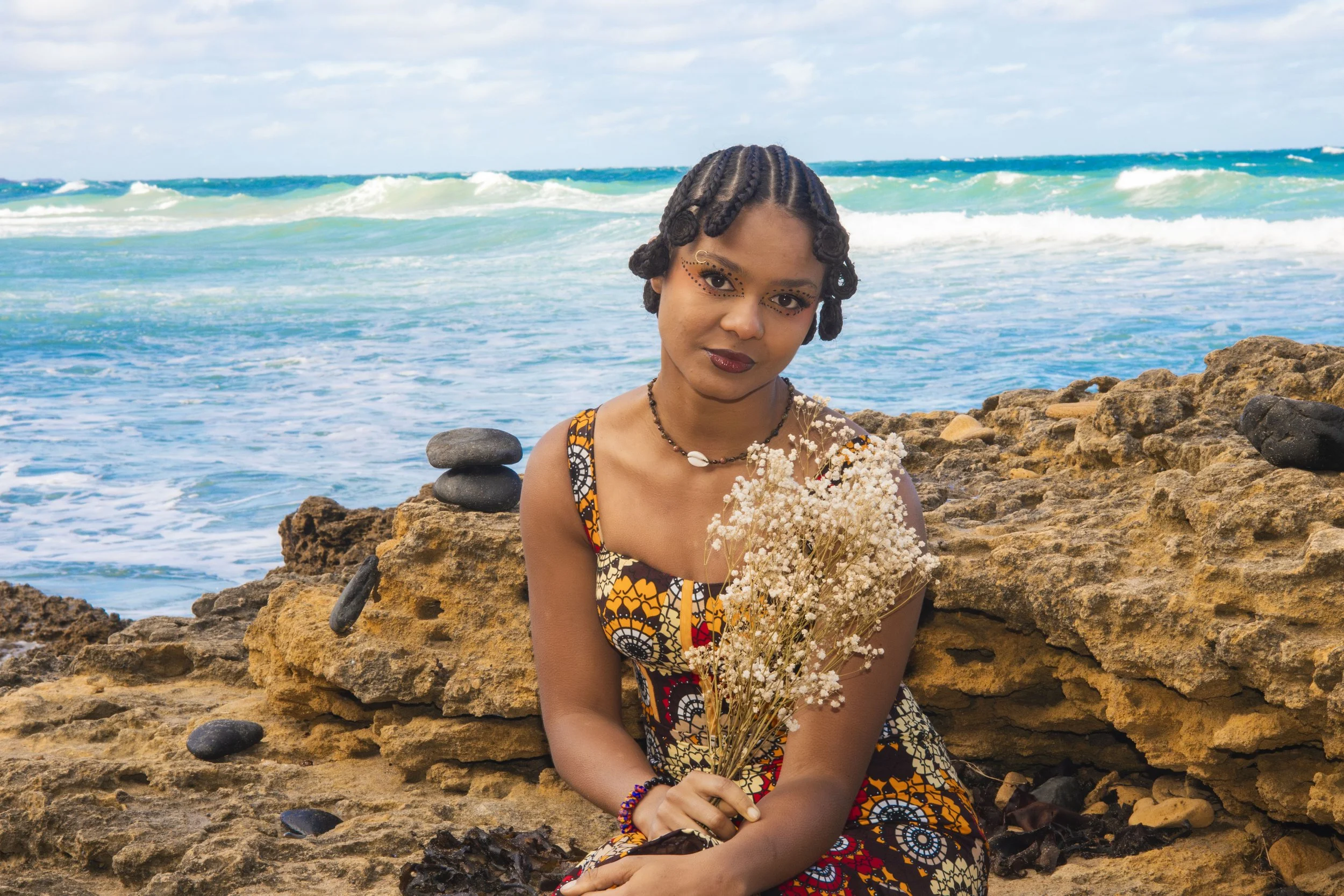 A woman with braided hair and artistic makeup sitting on rocks by the ocean, holding a bouquet of white flowers.
