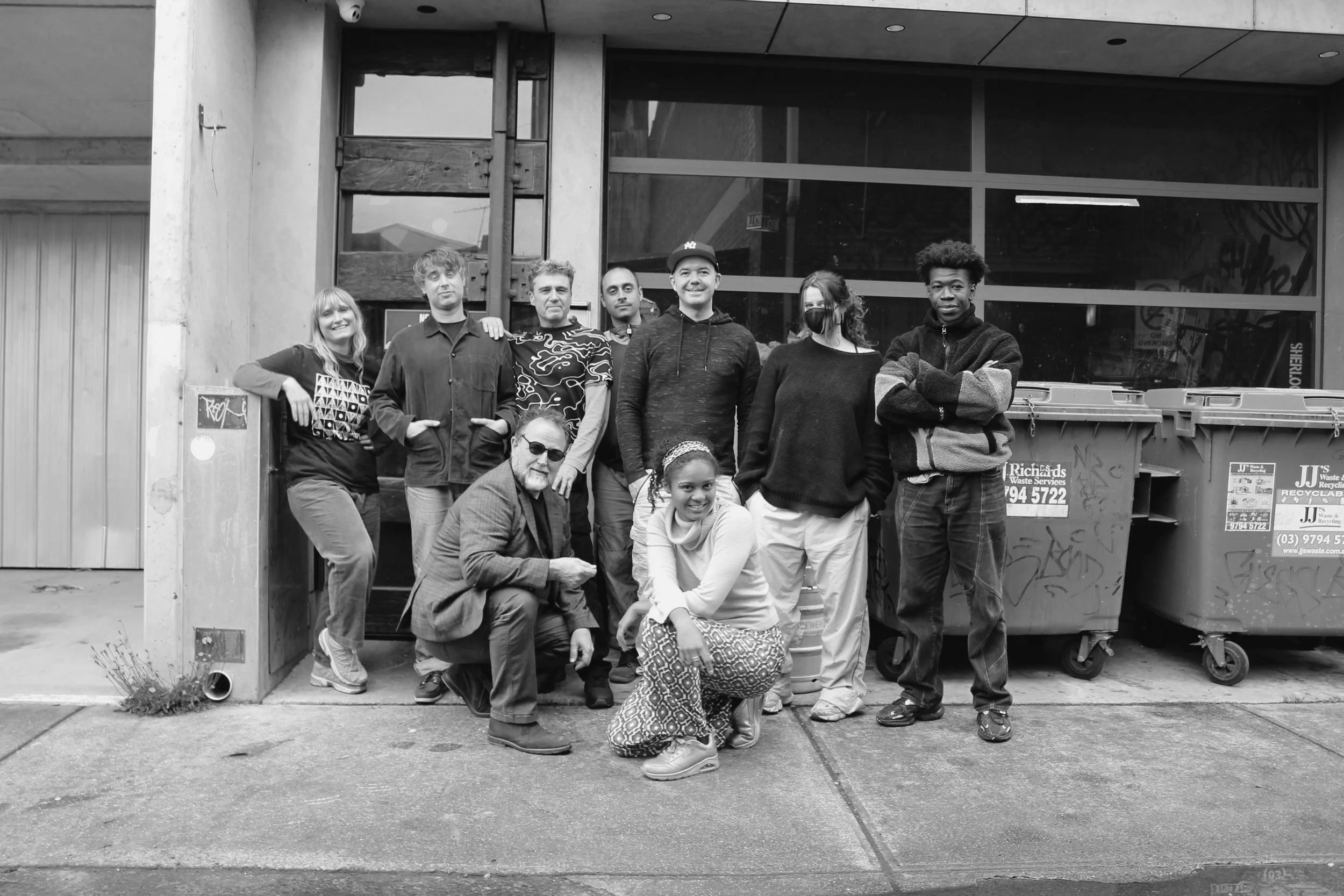 A group of nine diverse people, including men, women, and children, posing together outside an urban building with large glass windows and a concrete facade. Some are smiling and wearing casual clothing, with one woman wearing a mask.
