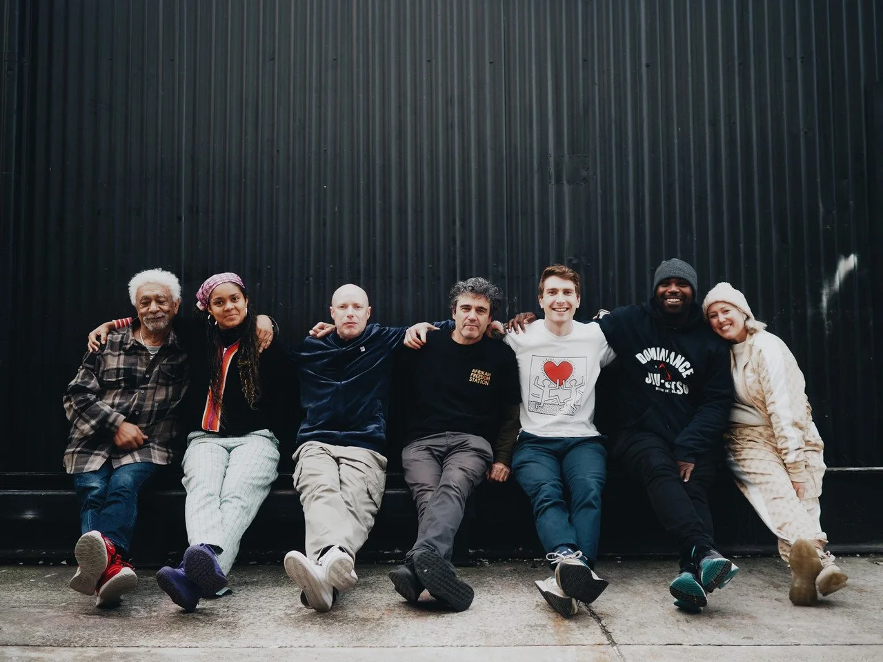 Group of seven diverse people sitting on a bench against a black wall, smiling and relaxed.