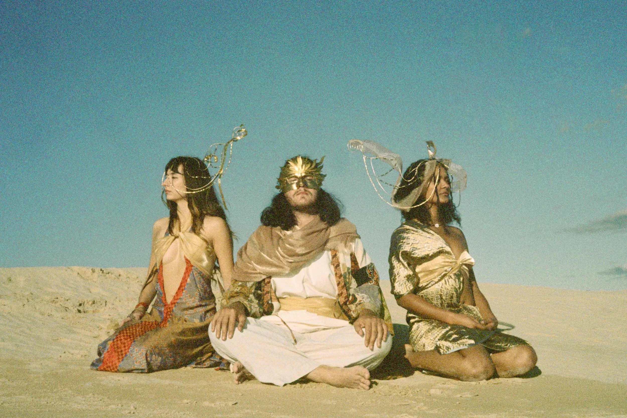 Three people sitting on the sand at the beach under a clear blue sky. The person in the middle is wearing a gold mask and a brown hooded sweatshirt, with two women on either side wearing shawls and large decorative headpieces.