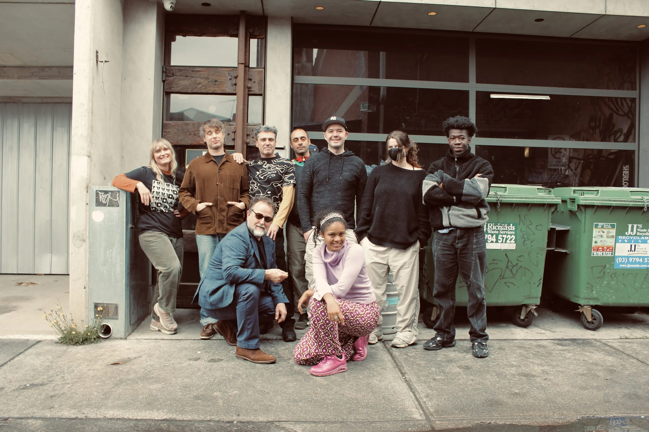 A diverse group of nine people, including a young girl, posing in front of a building with large windows. The group includes men and women of different ages and ethnicities, some wearing casual clothing, one with a face mask, and one man with sunglasses. There are two large green recycling bins nearby.