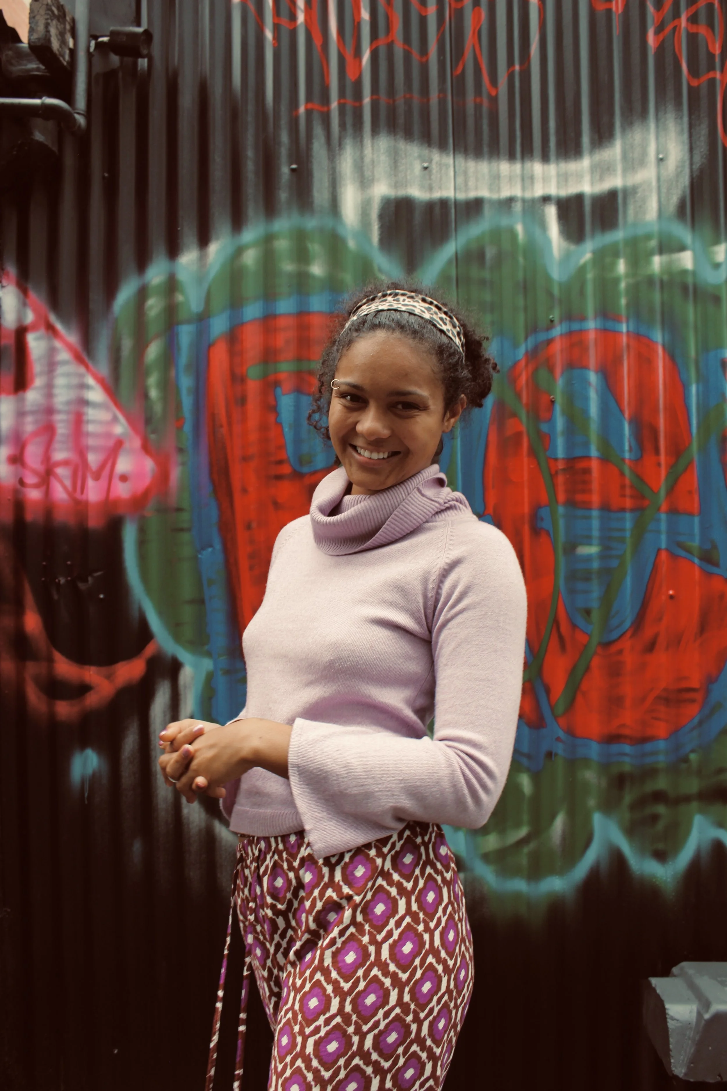 A woman wearing a light purple turtleneck sweater and patterned skirt, smiling, standing in front of a colorful mural on a corrugated metal wall.