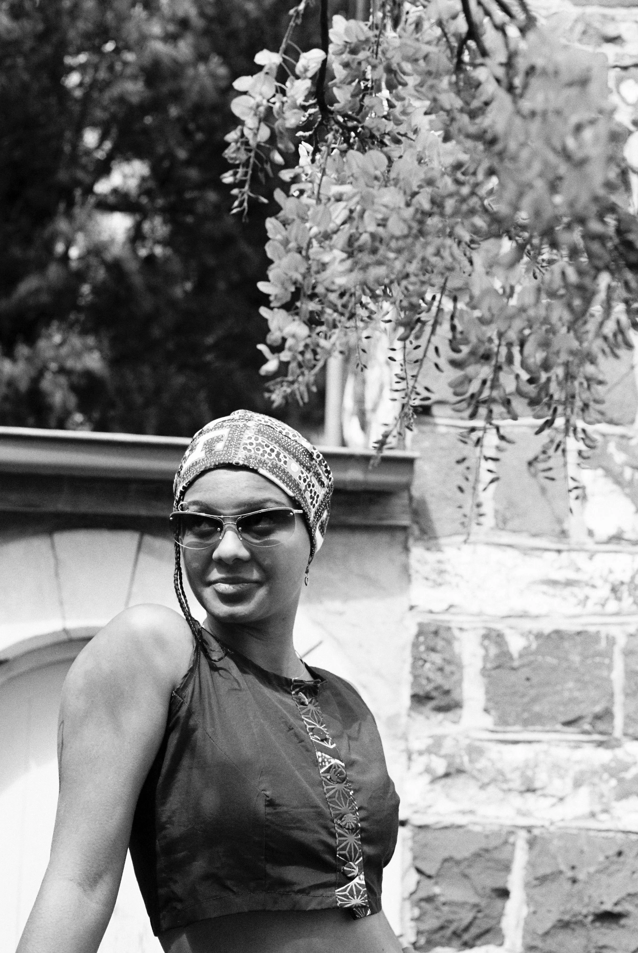 Black and white photo of a woman wearing sunglasses, a patterned headscarf, and a sleeveless top, standing outdoors in front of a brick wall and tree with hanging branches.