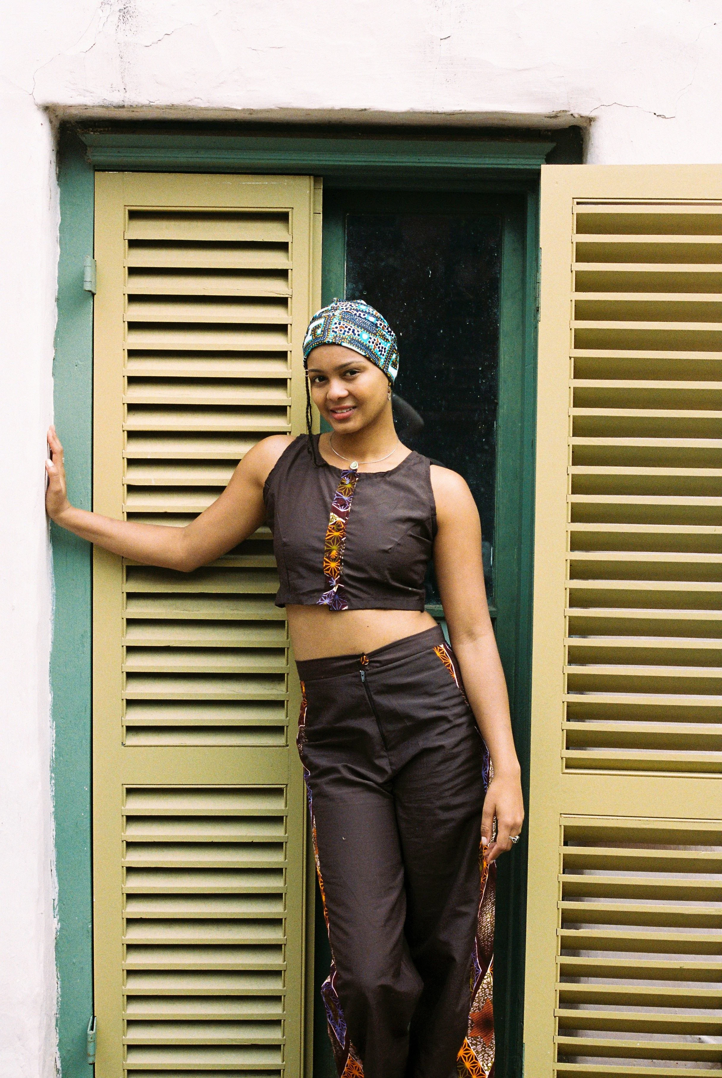A woman wearing a colorful headscarf and dark clothing is standing in front of an open window with yellow shutters, leaning against the wall and smiling.