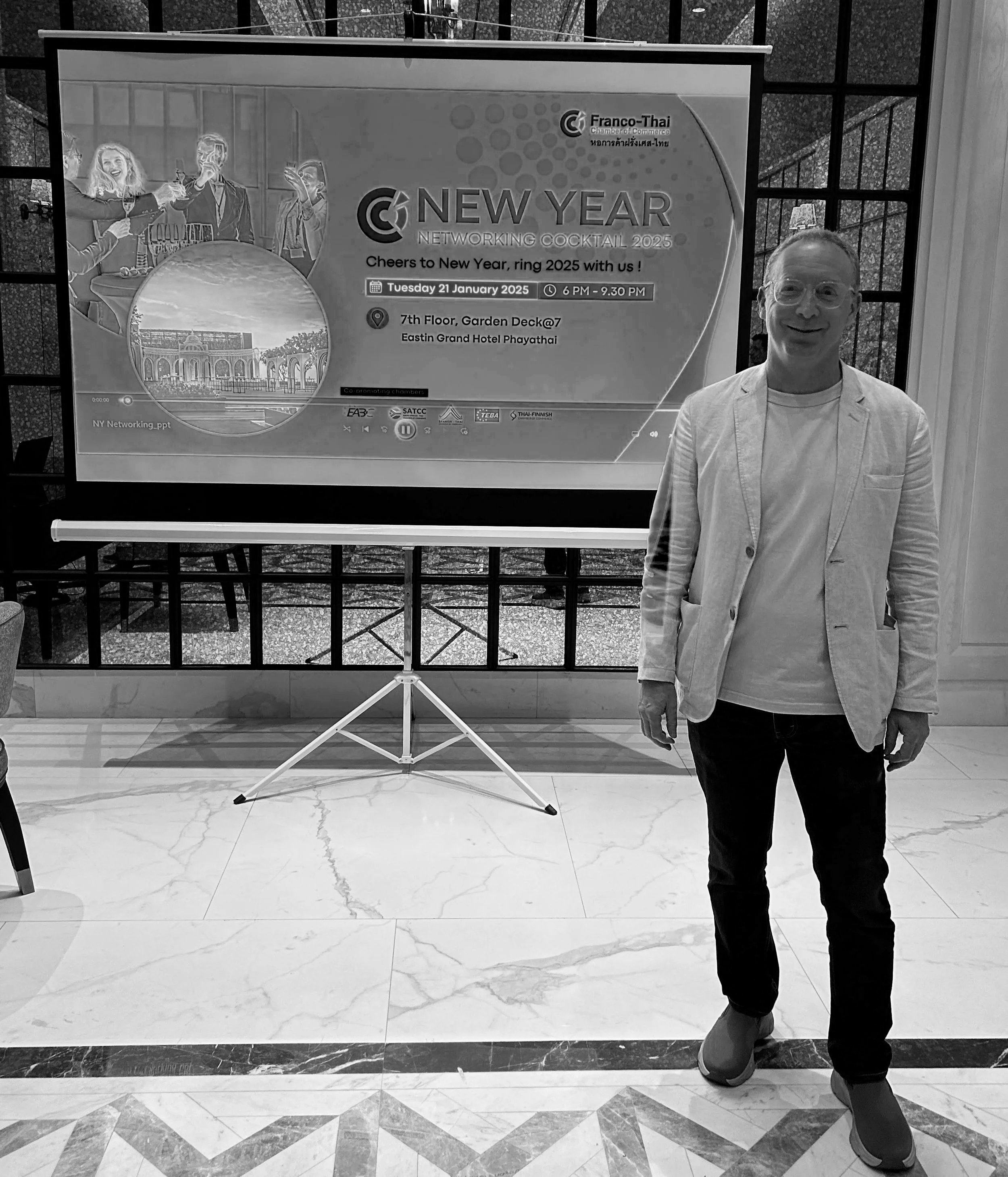 Un homme se tient à côté d'un écran de présentation affichant des informations sur un cocktail de réseautage du Nouvel An organisé par la Chambre de commerce franco-thaïlandaise, prévu pour le 21 janvier 2025 à l'hôtel Eastin Grand Phayathai.