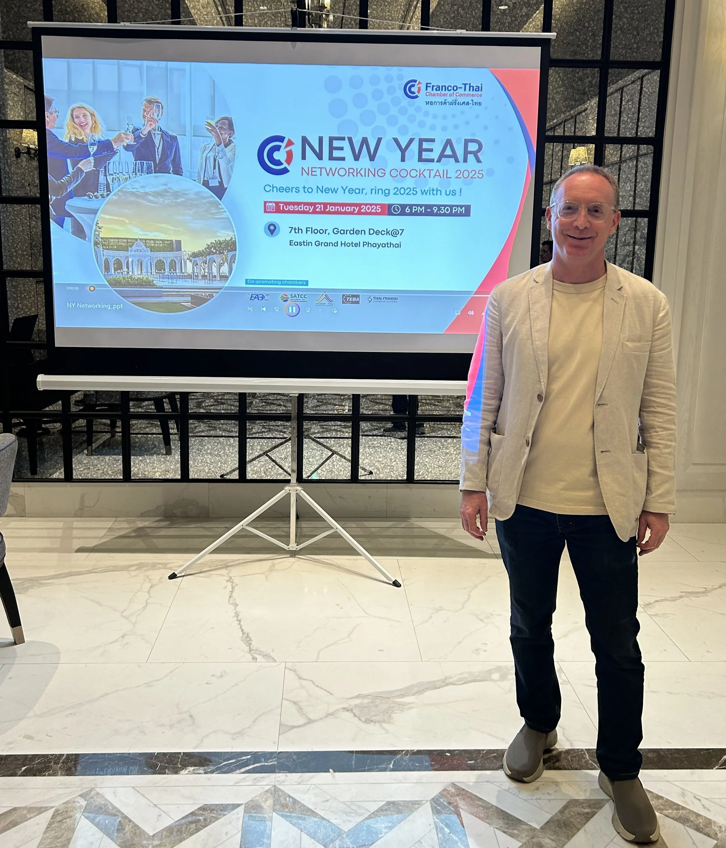 Un homme se tient à côté d'un écran de présentation affichant des informations sur un cocktail de réseautage du Nouvel An organisé par la Chambre de commerce franco-thaïlandaise, prévu pour le 21 janvier 2025 à l'hôtel Eastin Grand Phayathai.
