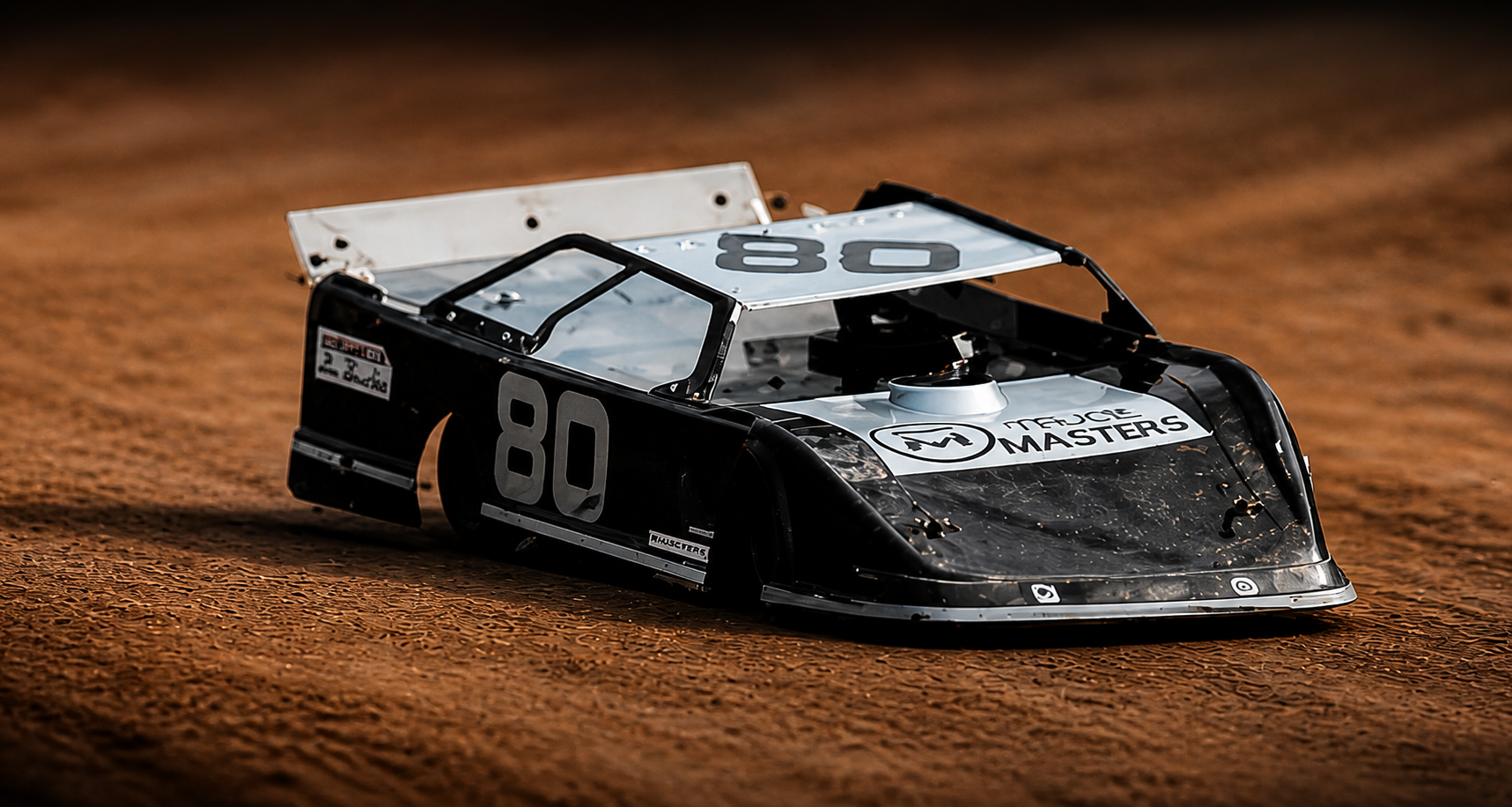 A black and silver slot car with the number 80 on its side and top, racing on a dirt track, featuring sponsorship logos including 'Mercize Masters'.