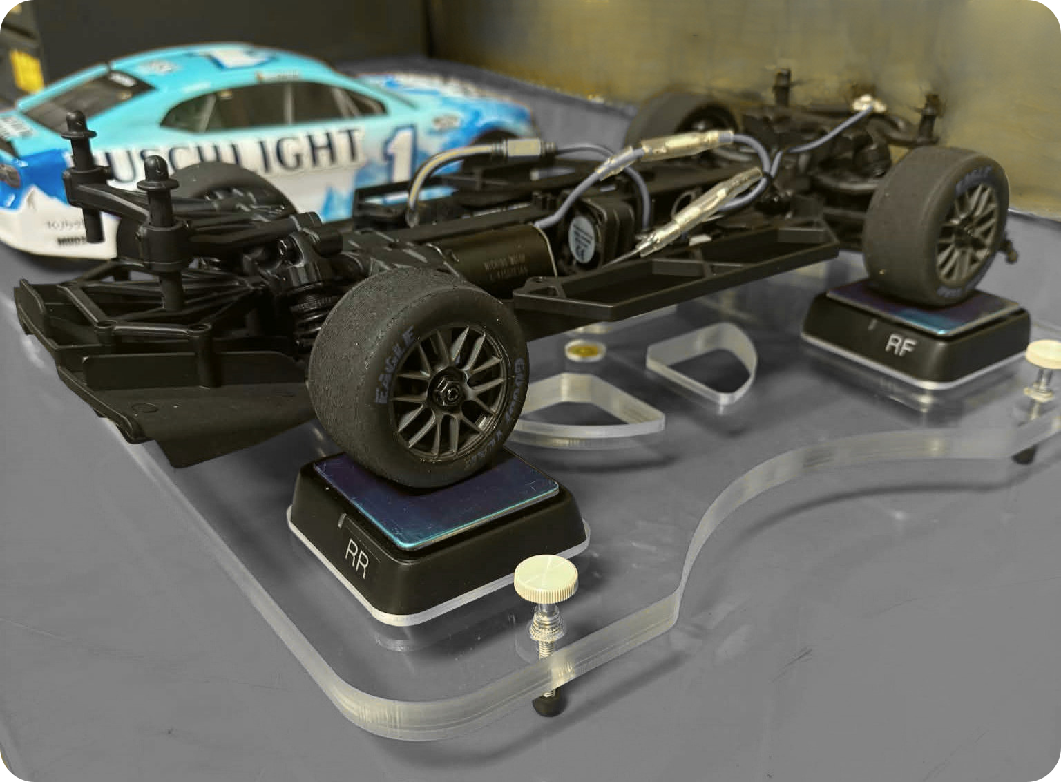 Close-up of a remote-controlled race car chassis with wheels, electronics, and suspension, placed on a display stand with labeled platforms.