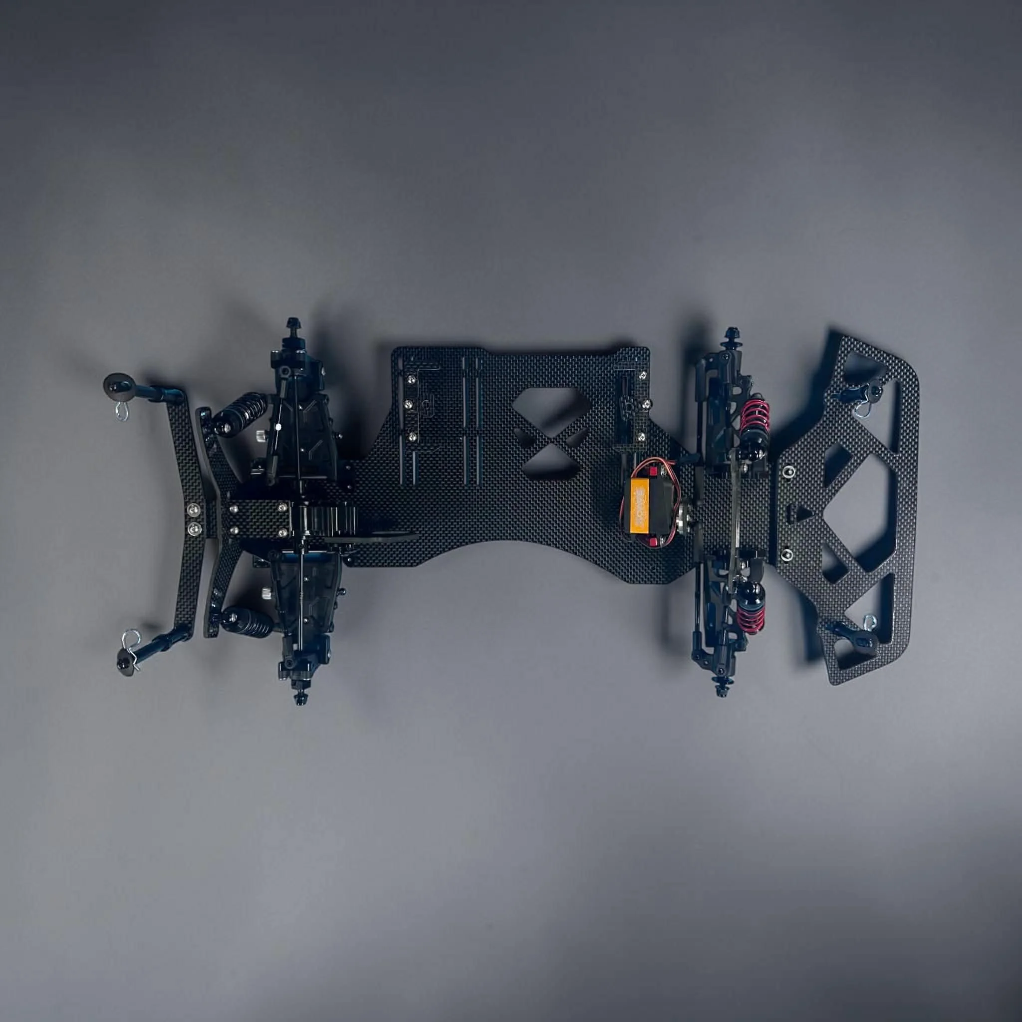 A partially assembled remote control car chassis with carbon fiber frame, suspension components, and electronic wiring visible on a gray background.