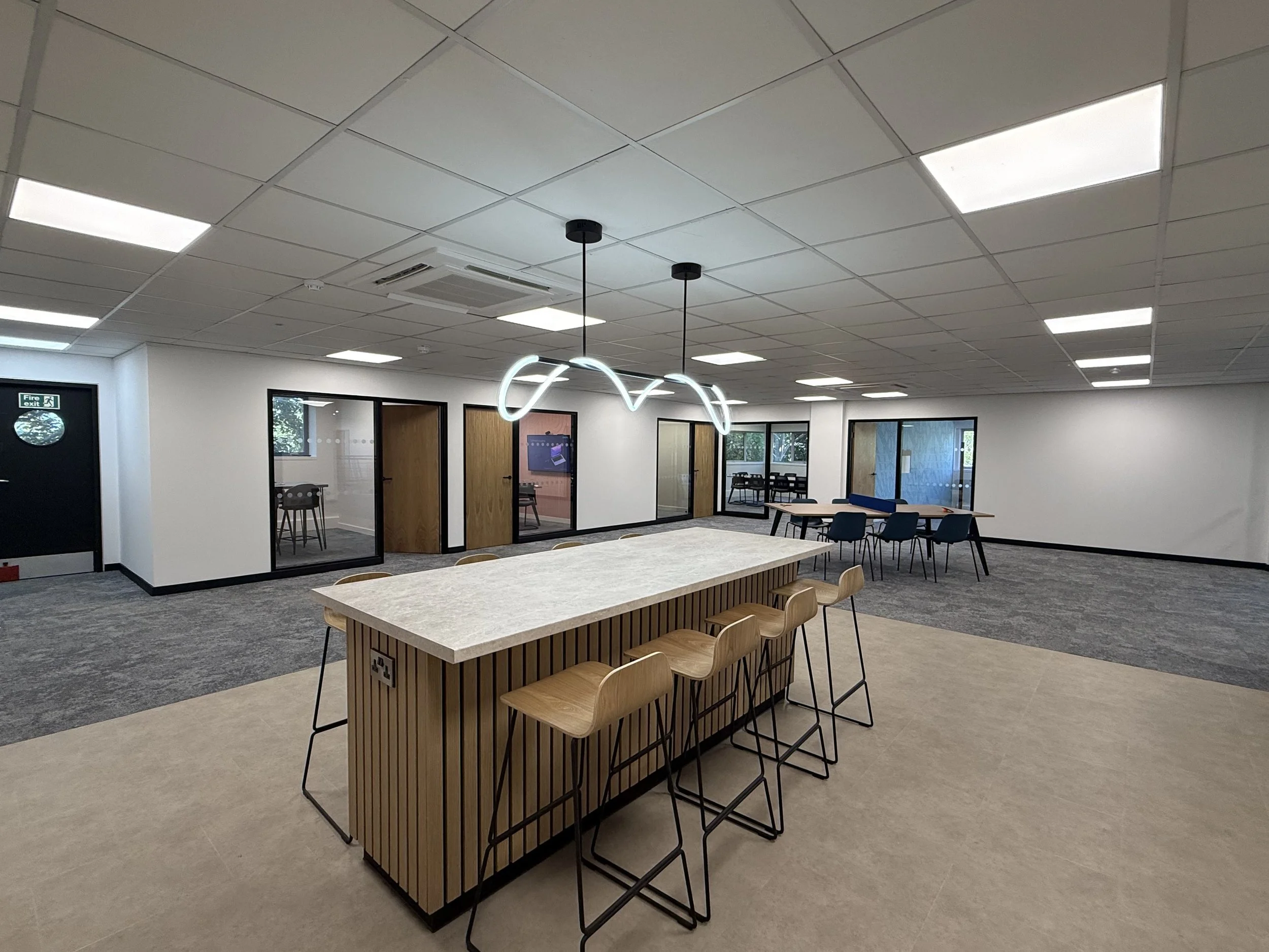 Modern office space with a large wooden communal table, bar-height chairs, and a high ceiling with fluorescent lights and a decorative hanging light fixture. There are glass partitioned rooms and windows.