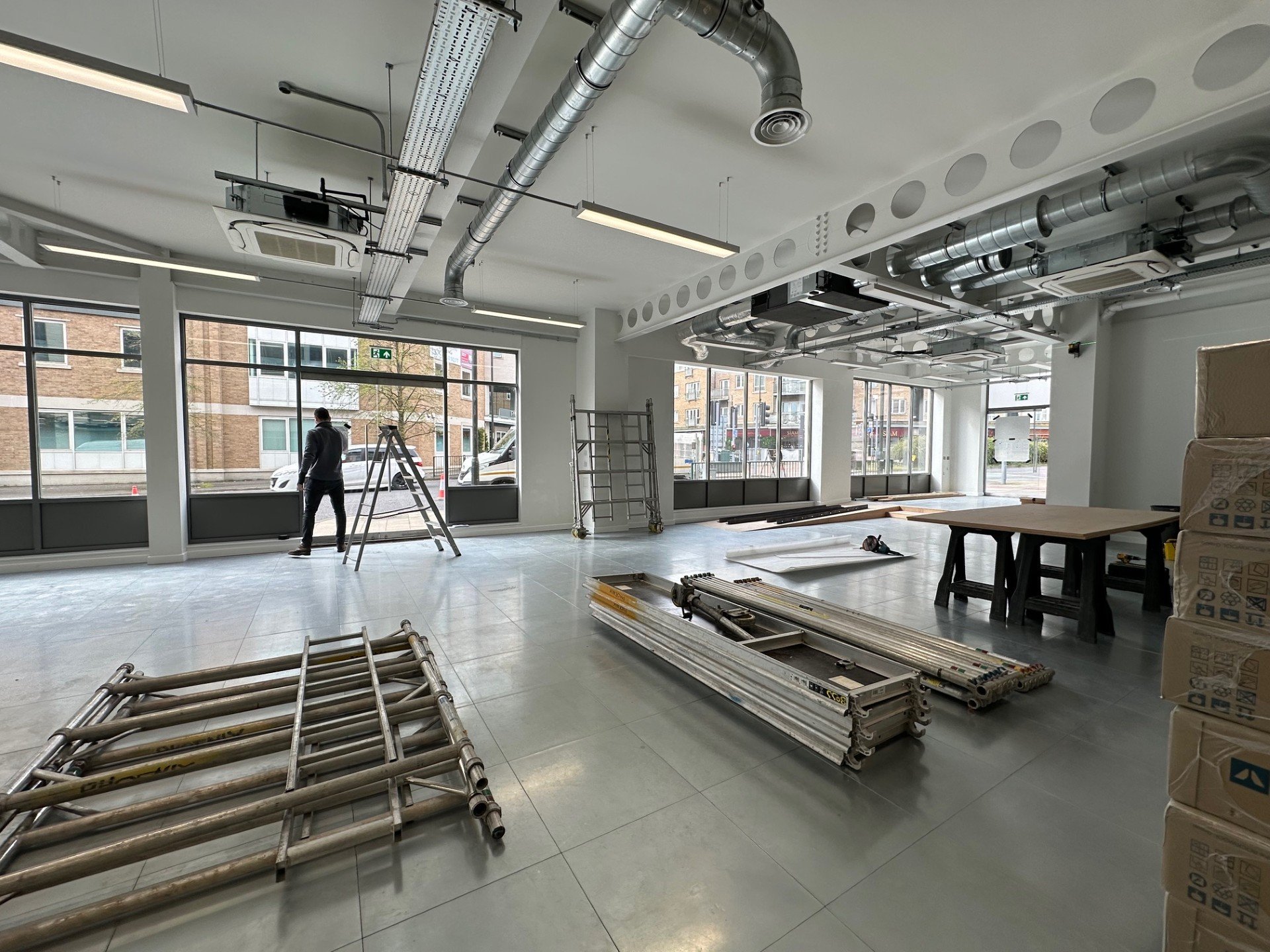 Interior of a commercial space under construction with large glass windows, metal scaffolding and orange cones outside, construction equipment and tools inside, and trucks visible through the windows.