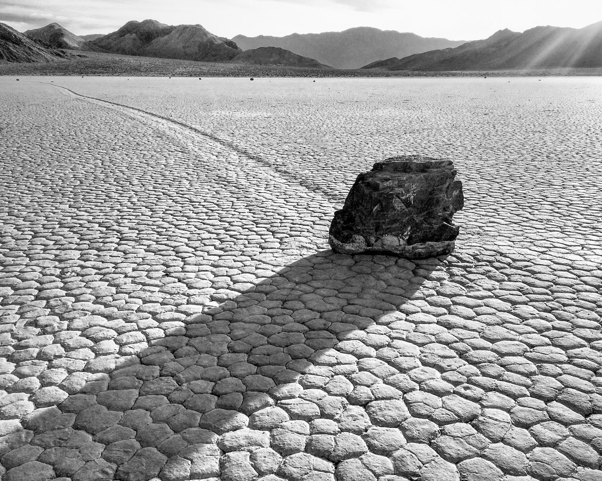 Black and white photo of a boulder on a cracked mud lake bed casting a long shadow with a trail curving into the distance.