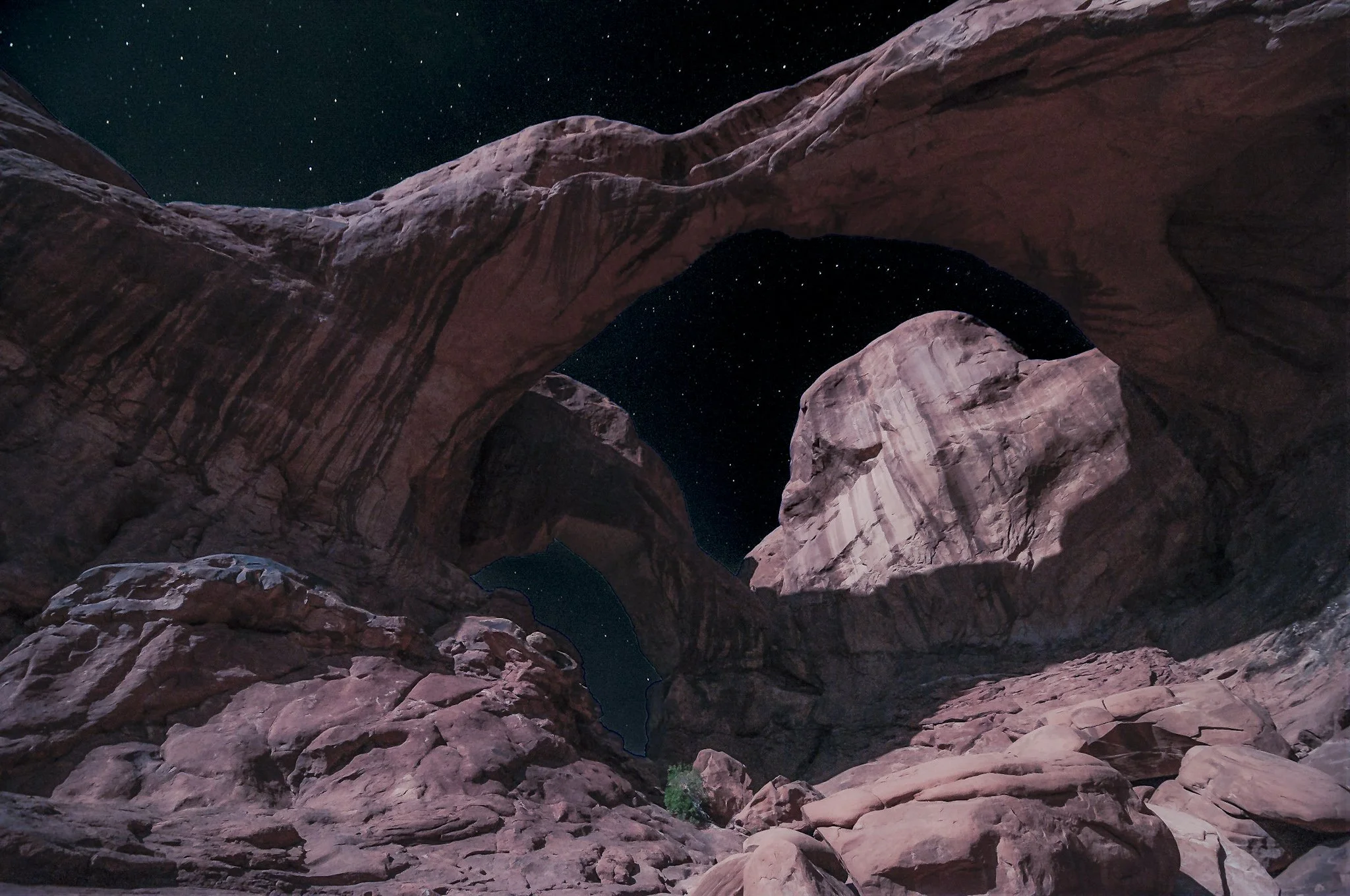 A night landscape photograph of the Double Arch in Arches National Park, illuminated by bright moonlight with stars visible through the openings of the arches.