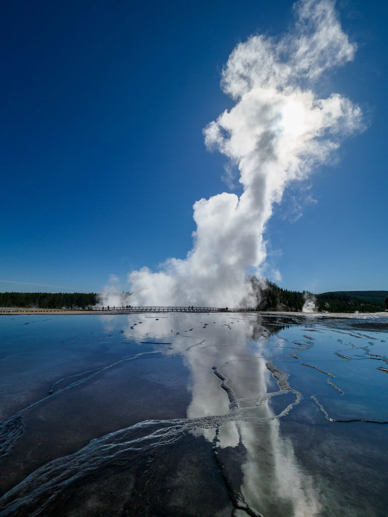 A towering white steam plume from a Yellowstone geyser reflects perfectly in a mineral-lined pool under a bright blue sky.