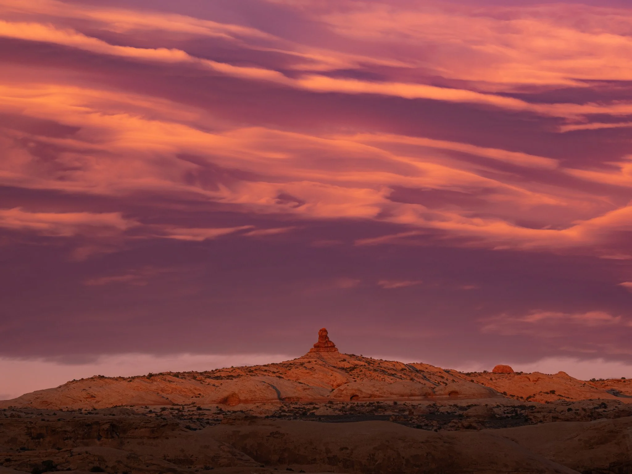 A fiery sunset with dramatic magenta and orange clouds sweeping over the desert horizon and petrified dunes in Arches National Park.