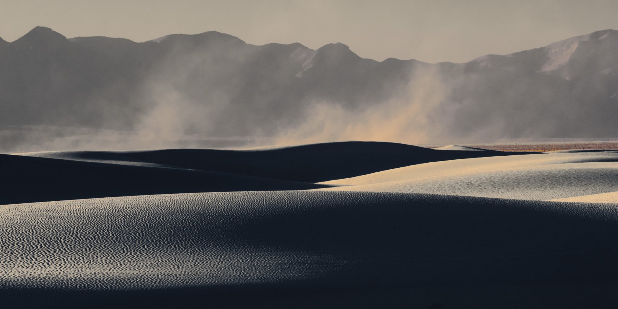 Swirling dust devil rising over sweeping white gypsum sand dunes and dark mountain silhouettes at White Sands National Park.