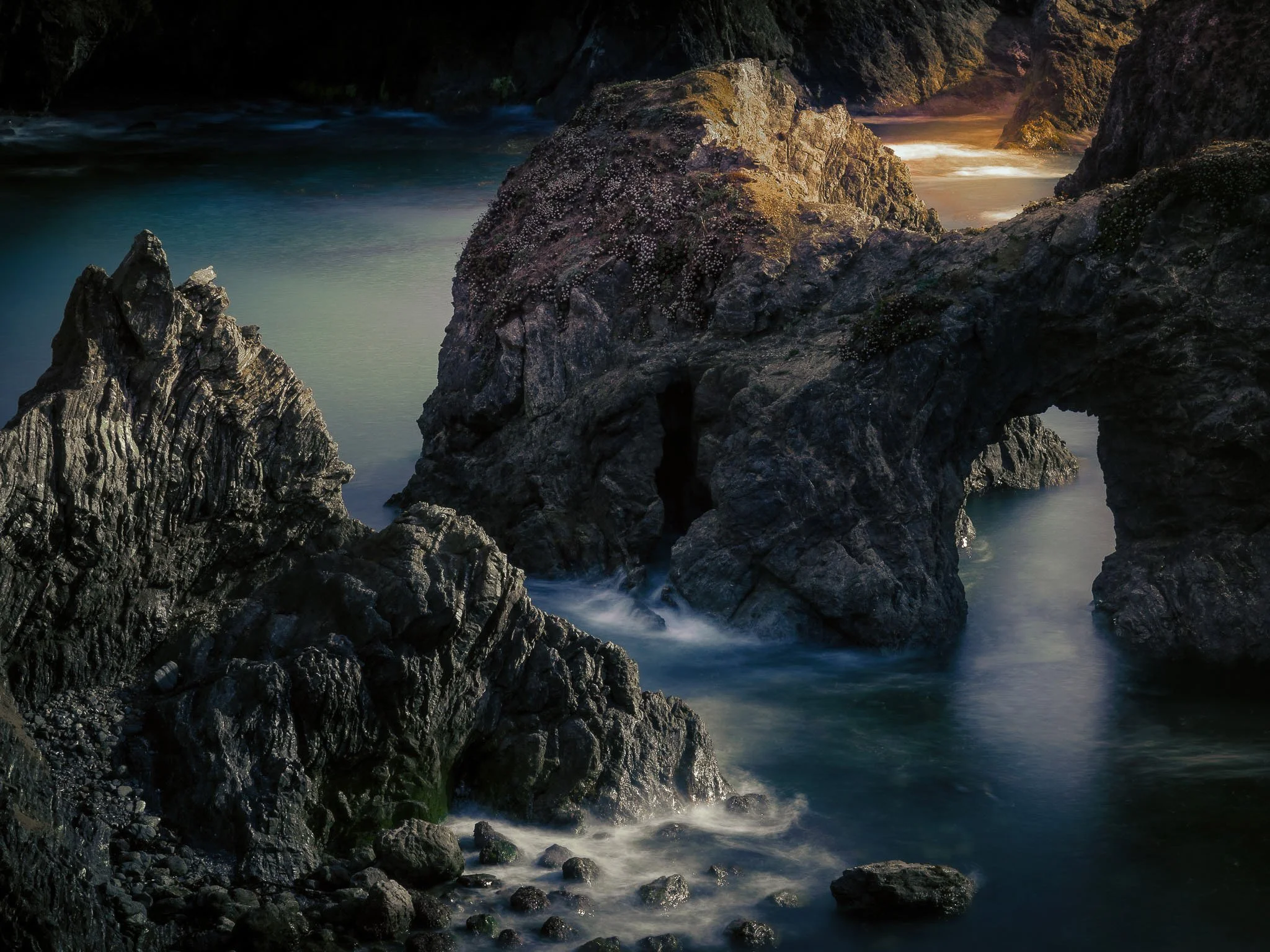 A long-exposure seascape of rugged sea stacks and a natural stone arch along the windy Mendocino California coast with misty water.