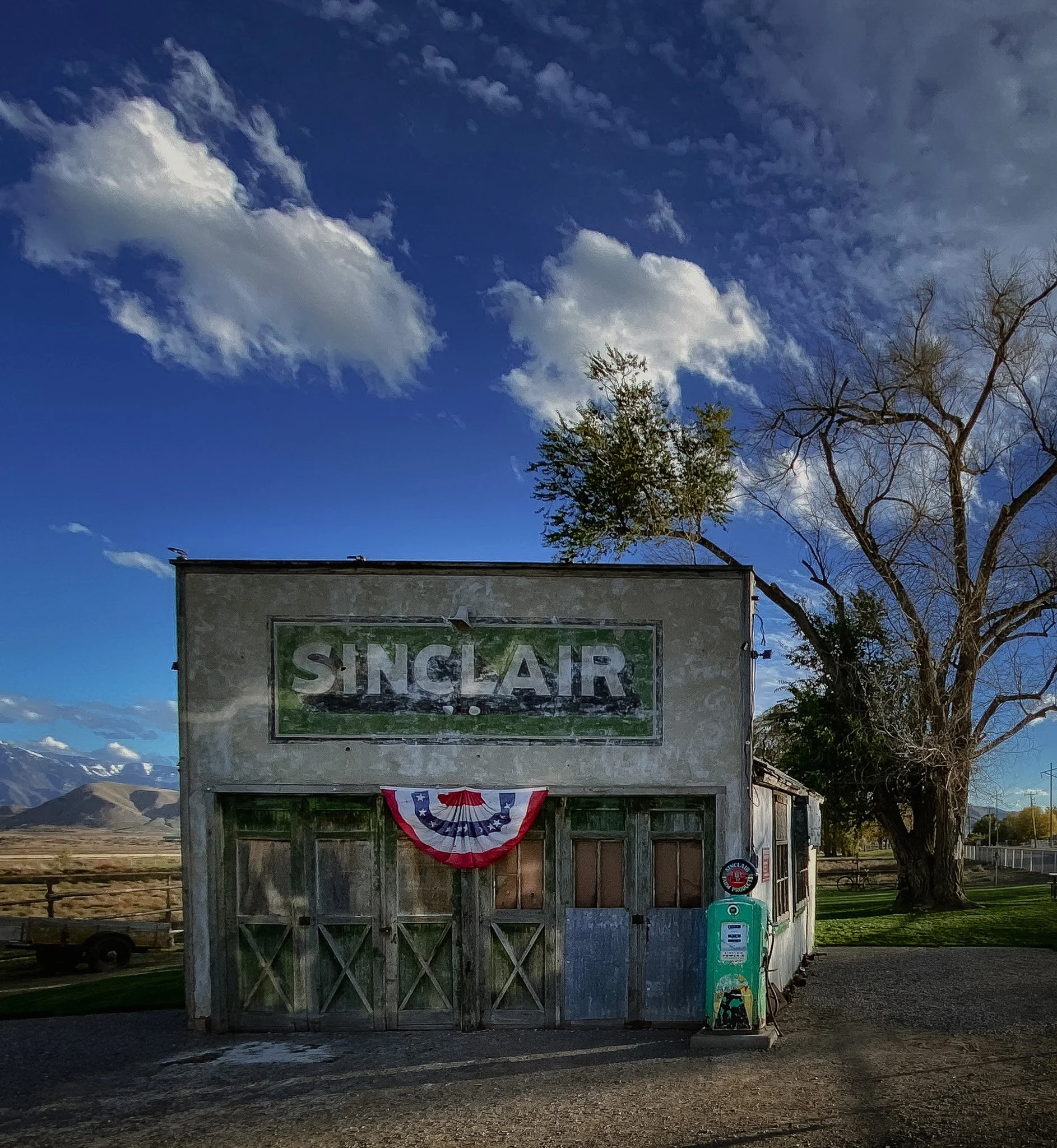 Historic abandoned Sinclair gas station with weathered wood doors, vintage green pump, and patriotic bunting under a blue sky in Utah.