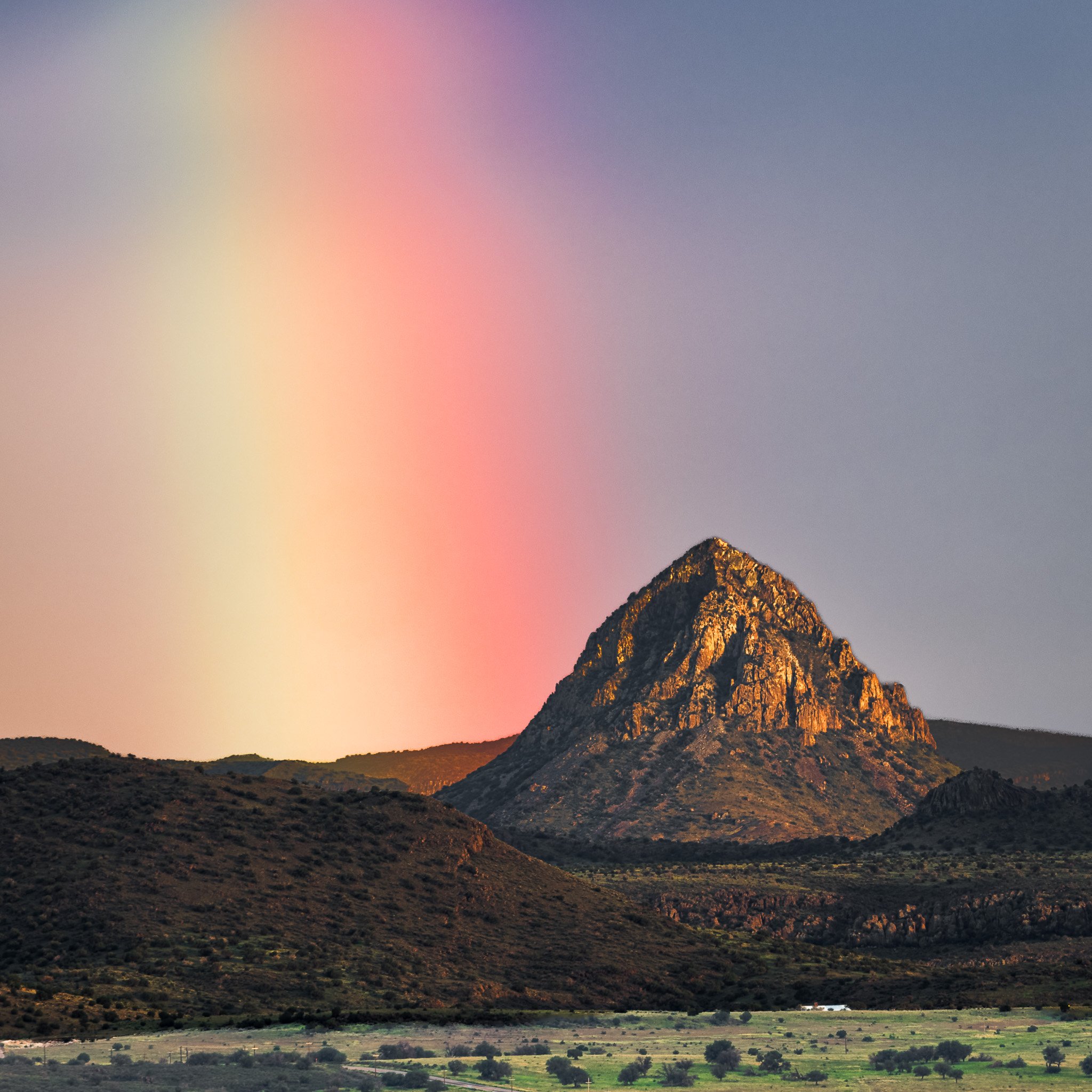 A colorful rainbow beam shines brightly in the sky next to the rugged, sunlit peak of Mitre Peak in West Texas.