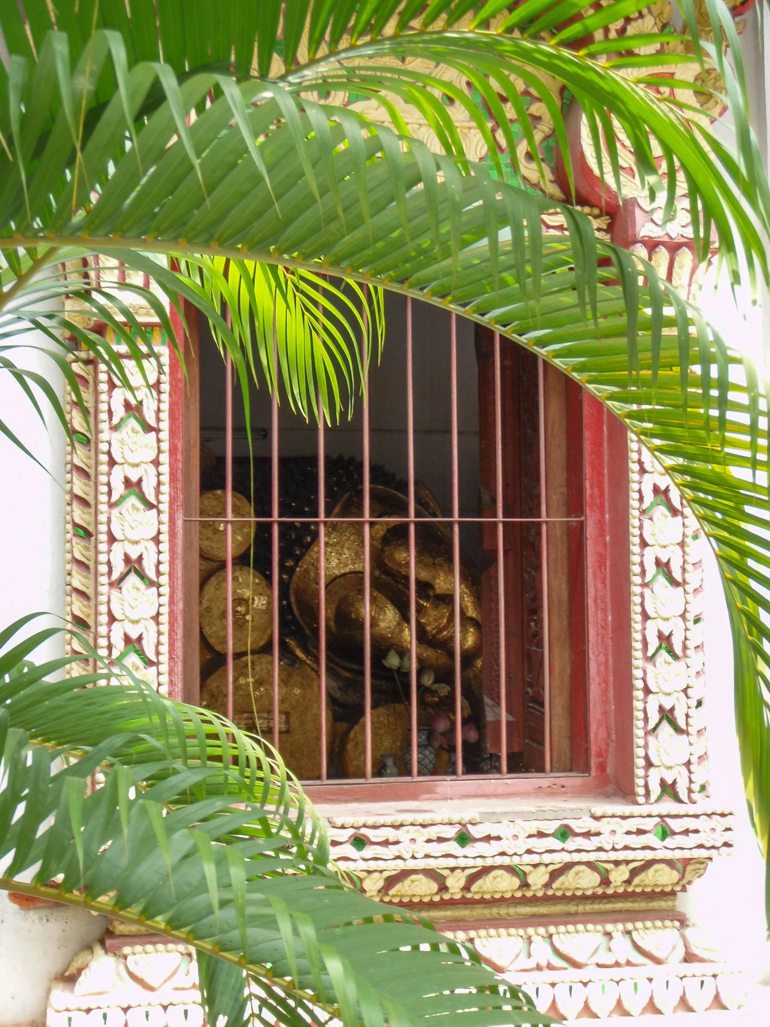 A golden Reclining Buddha statue seen through a window with bars and green palm leaves in Chiang Mai.