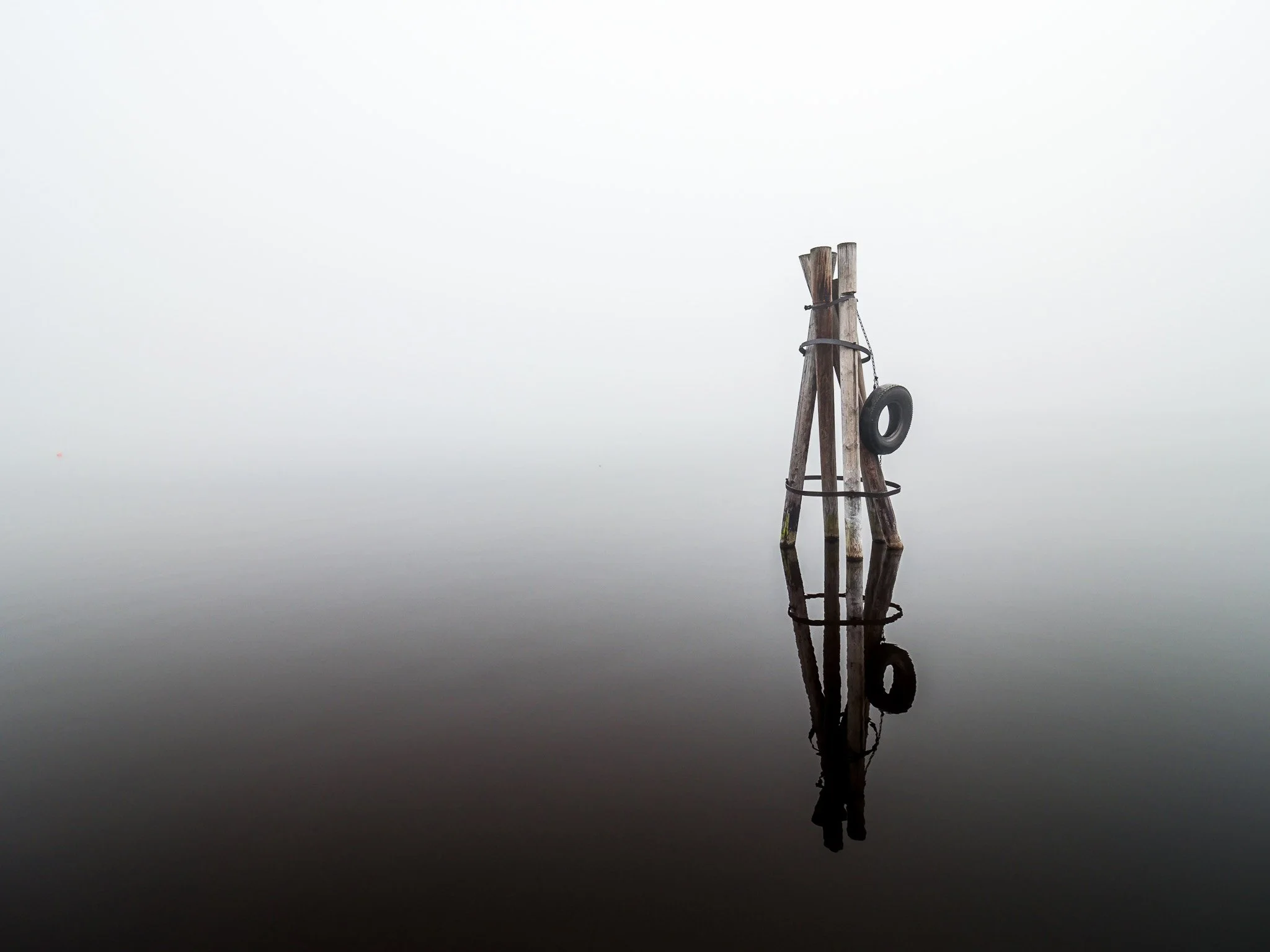 A minimalist high key photograph of a wooden pylon assembly with a single tire hanging from it, reflecting perfectly in the dark glassy water. The dense fog dissolves the background entirely, suggesting immense depth and isolation.