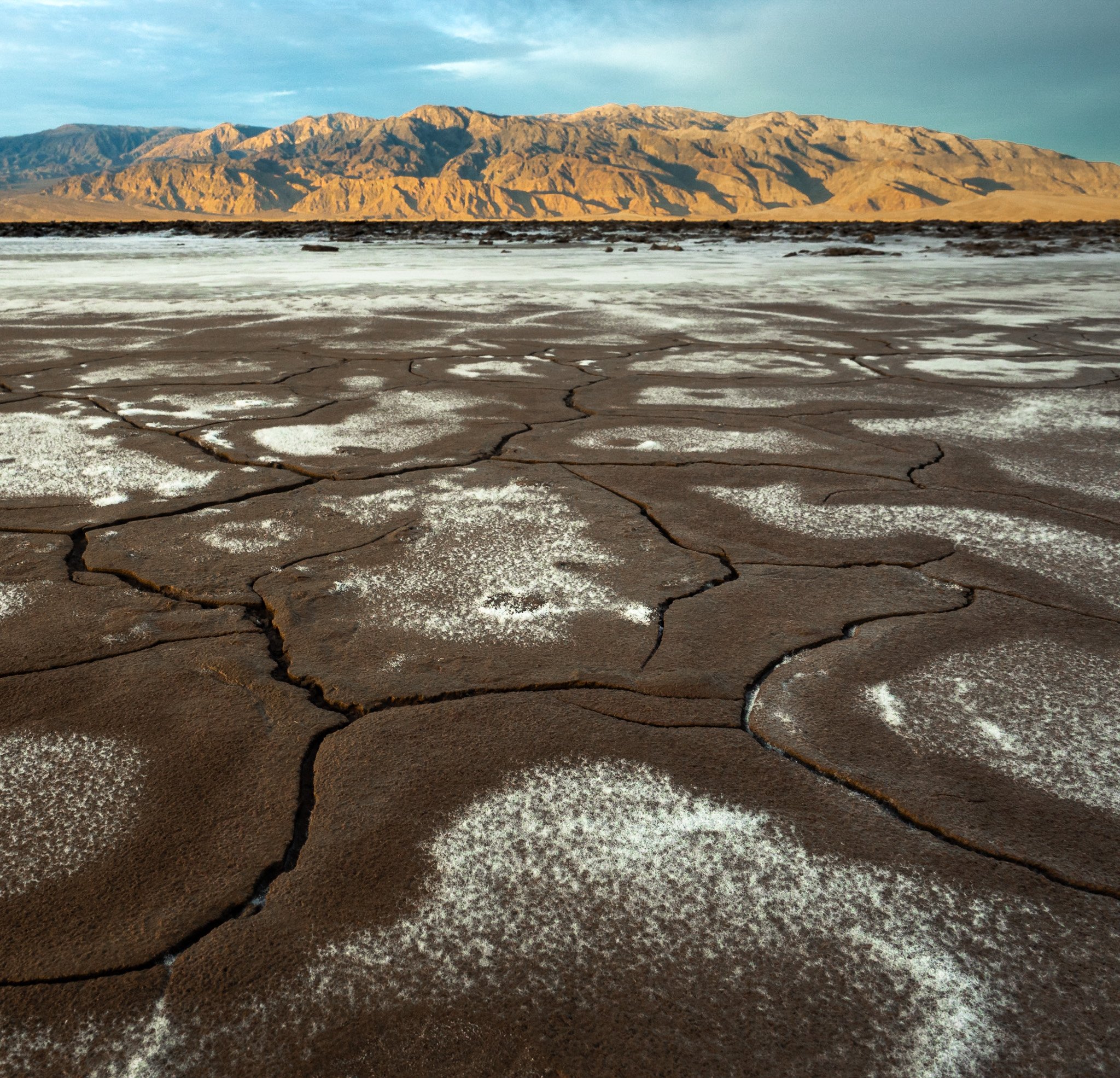 Sunbaked dark mud cracks filled with white salt deposits lead toward golden mountains under a blue sky in Death Valley National Park.