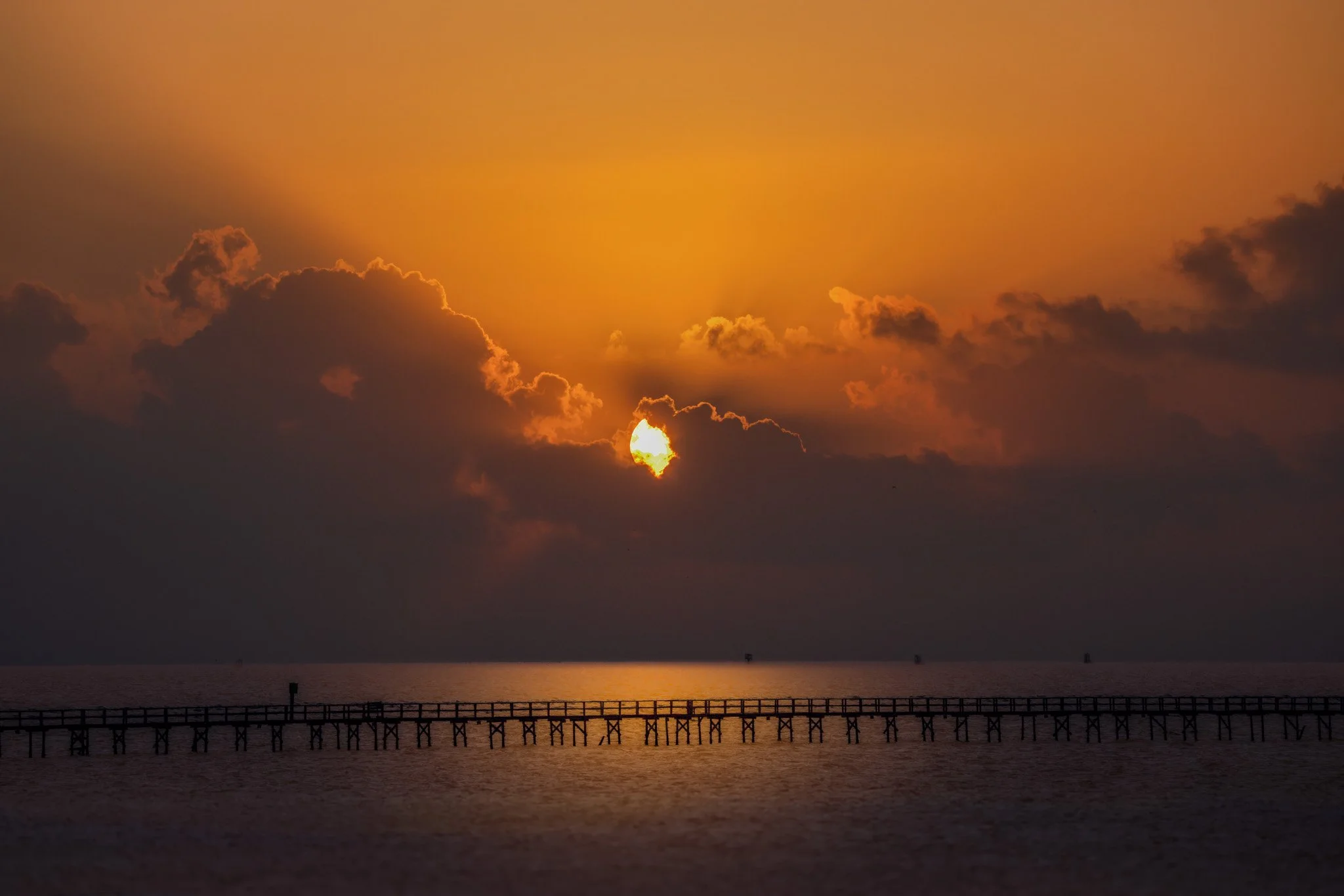 Golden sunrise breaking through clouds over a long fishing pier on Aransas Bay in Rockport, Texas, viewed from a waterfront deck.