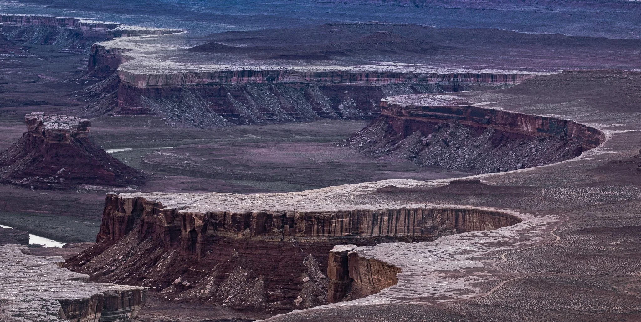 A wide winter landscape view of the deep rock benches and canyon layers of Canyonlands National Park, with the White Rim Road visible in the lower corner under overcast skies.