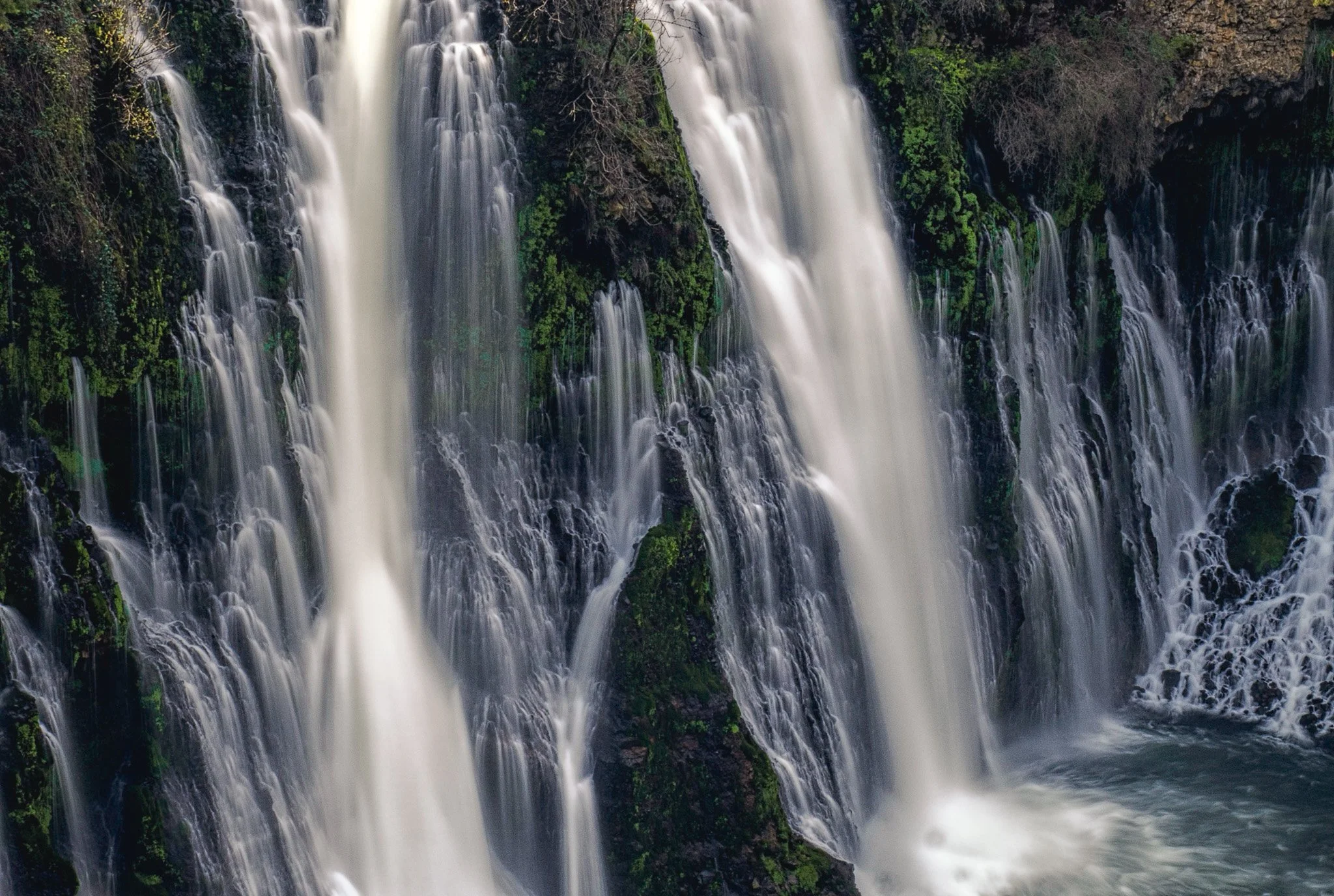 A long exposure photograph capturing the silky, rushing water of Burney Falls spilling over green, mossy volcanic rock.