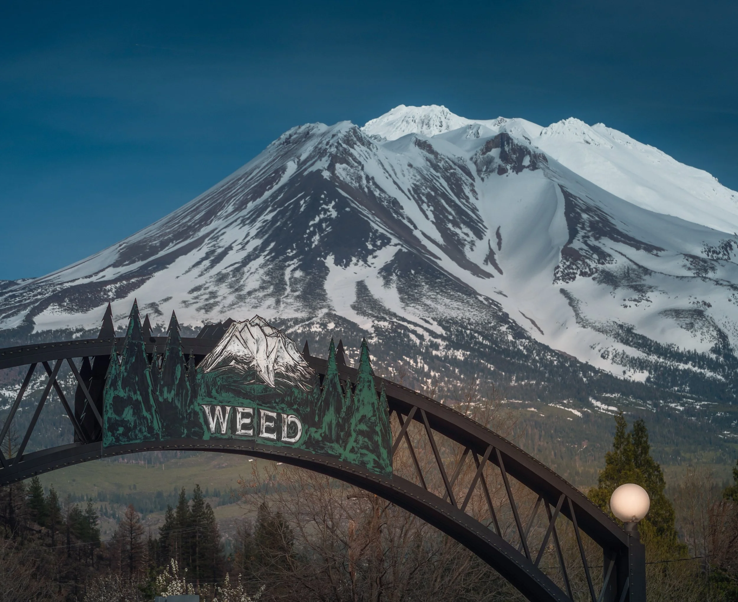Photo of a large metal truss archway. At the top center of the arch is a stylized sign,"Weed" and a small painted illustration of  Mt. Shasta, in the foreground. Behind, and towering over the arch is the massive, sacred and snow covered Mt. Shasta