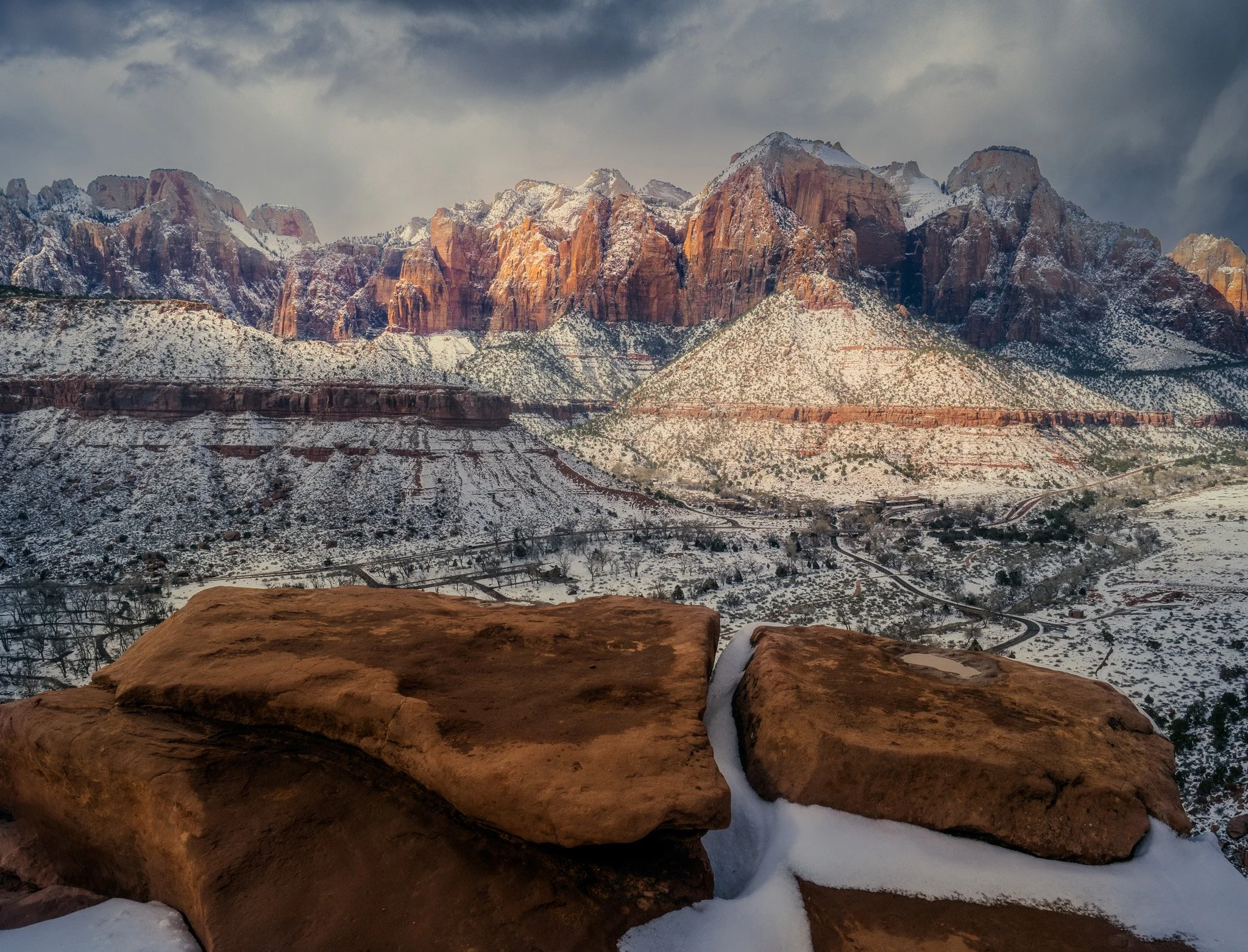 Red sandstone rocks with a snow filled gap in foreground, looking toward sunlit snow-capped cliffs in Zion National Park under storm skies.