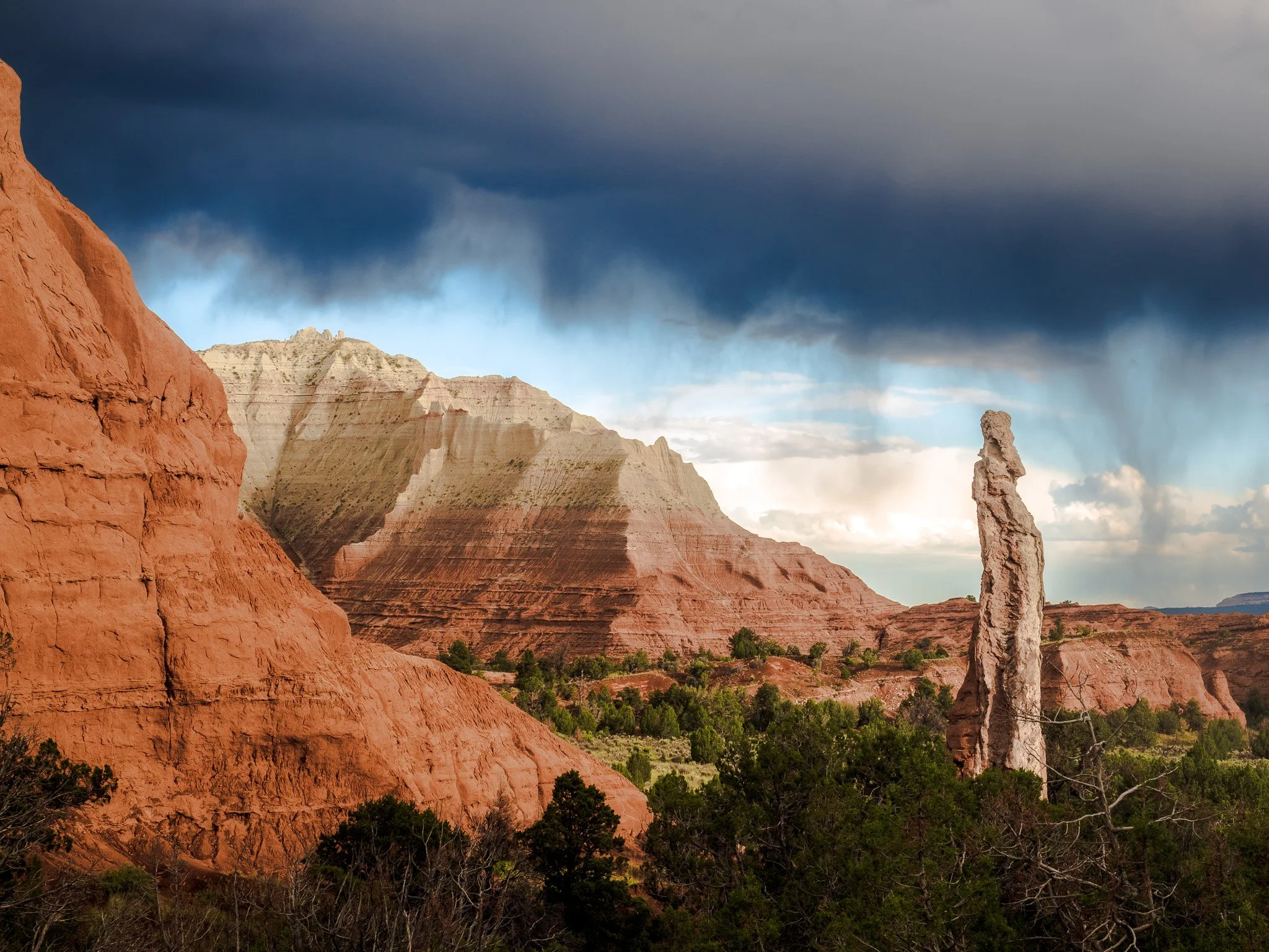 A tall sandstone spire stands against a backdrop of colorful desert cliffs and a localized rainstorm in Kodachrome Basin State Park, southern Utah