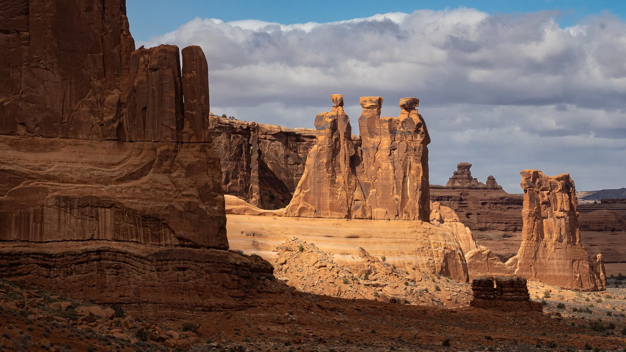A landscape photograph of the Three Gossips rock formation in Arches National Park, Utah, under a cloudy sky.