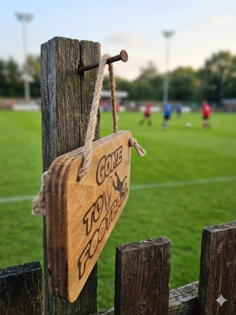 A close-up, perspective shot of a rustic wooden sign on a fence overlooking an active football field. The sign, reading I'd rather be at Football.jpg