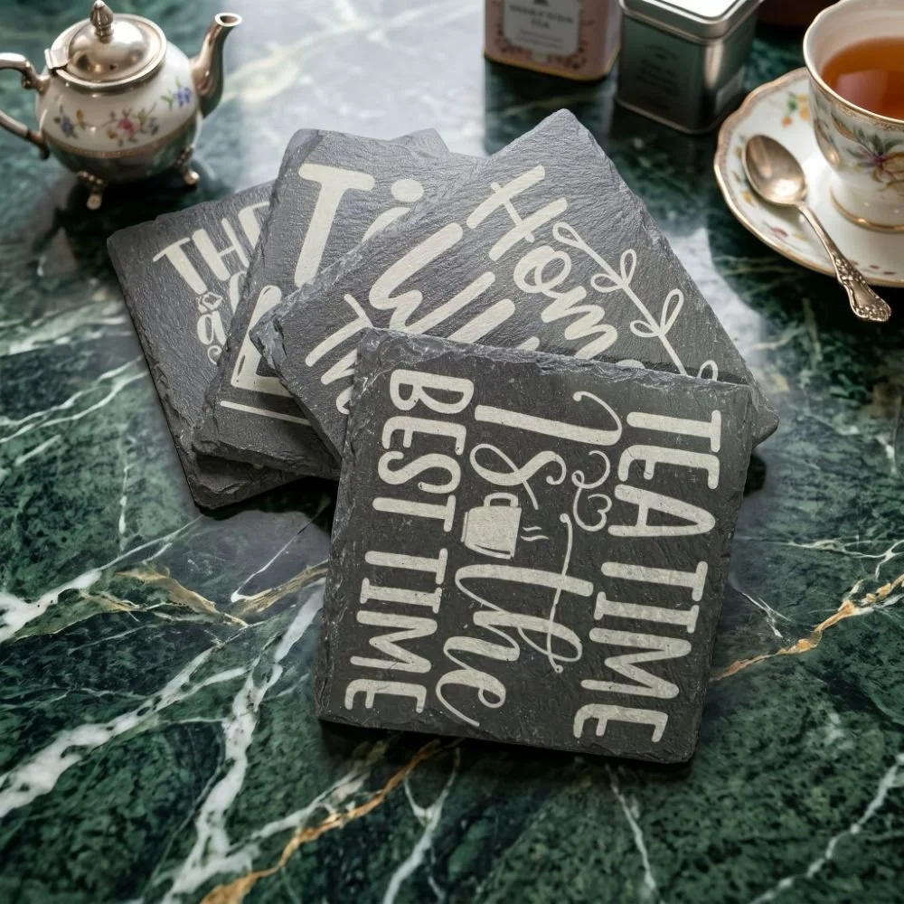 A stack of overlapping square dark grey slate coasters featuring laser-engraved white tea quotes,.jpeg