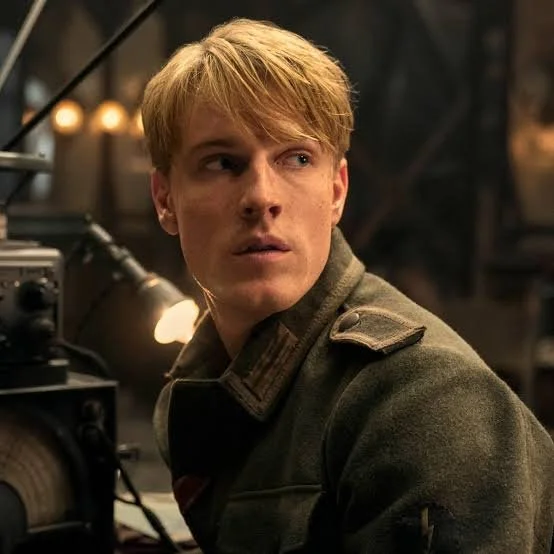 A young man with blond hair looking to his left in a dimly lit room with equipment and warm lighting.