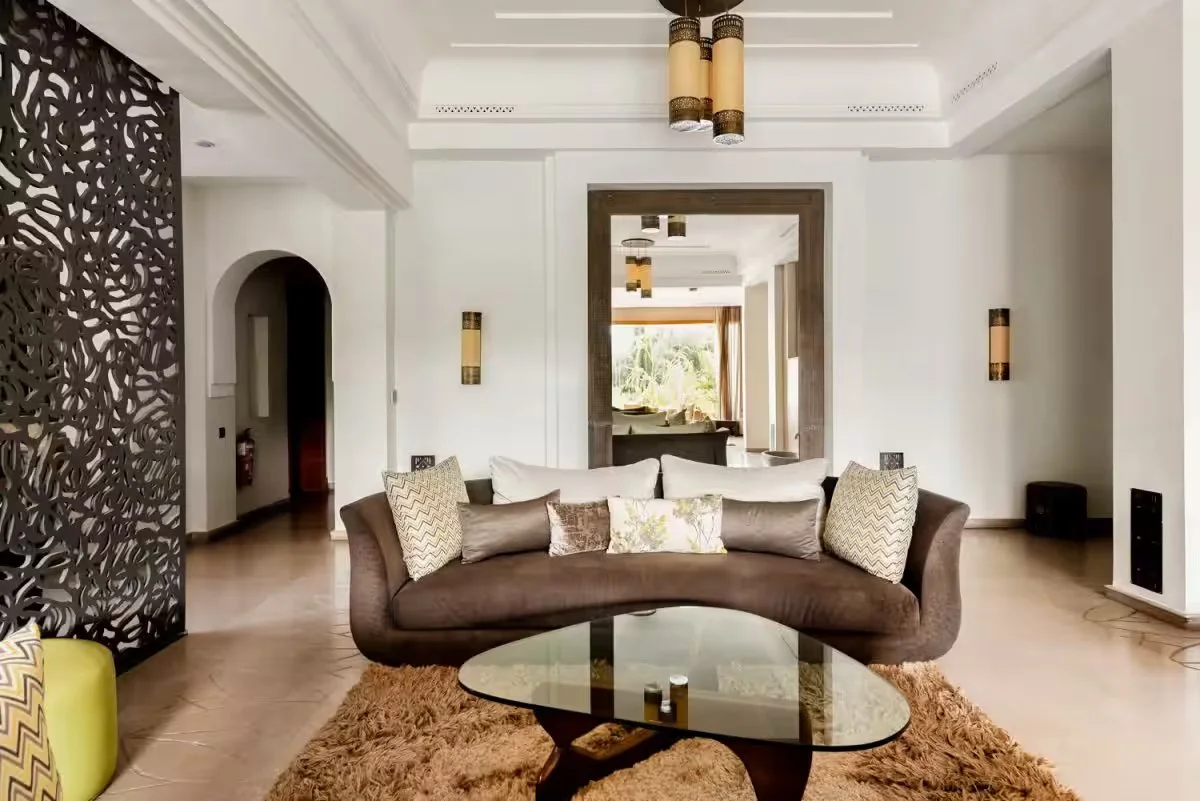 Salon moderne avec canapé marron, plusieurs coussins, table basse en verre, tapis en laine, murs blancs, décorations en bois, éclairage encastré, porte en bois menant à une autre pièce avec vue sur jardin Retraite pour femmes a marrakech au maroc